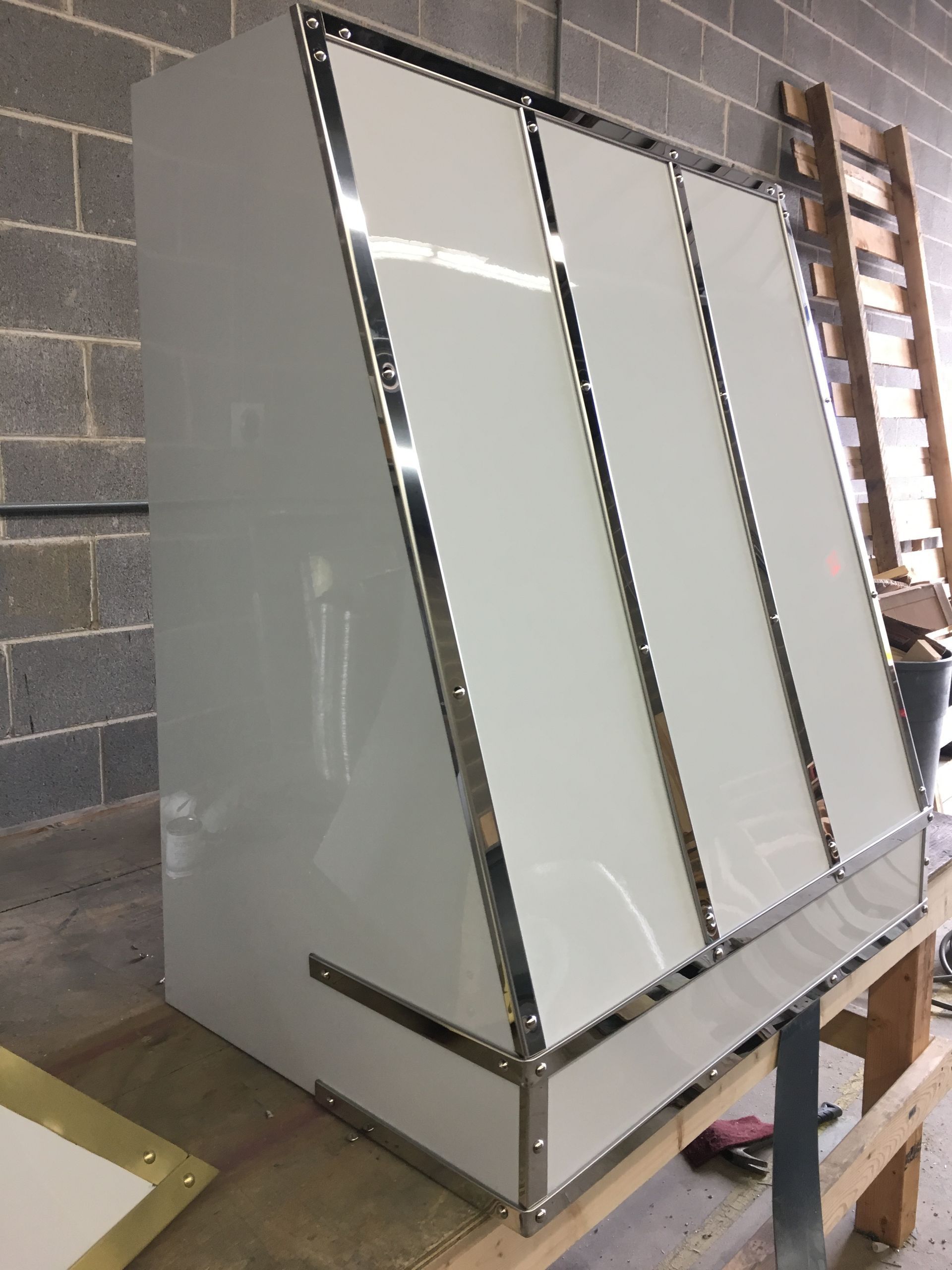 Large white metal cabinet or enclosure with angled front panels, sitting on a wooden workbench in a workshop.