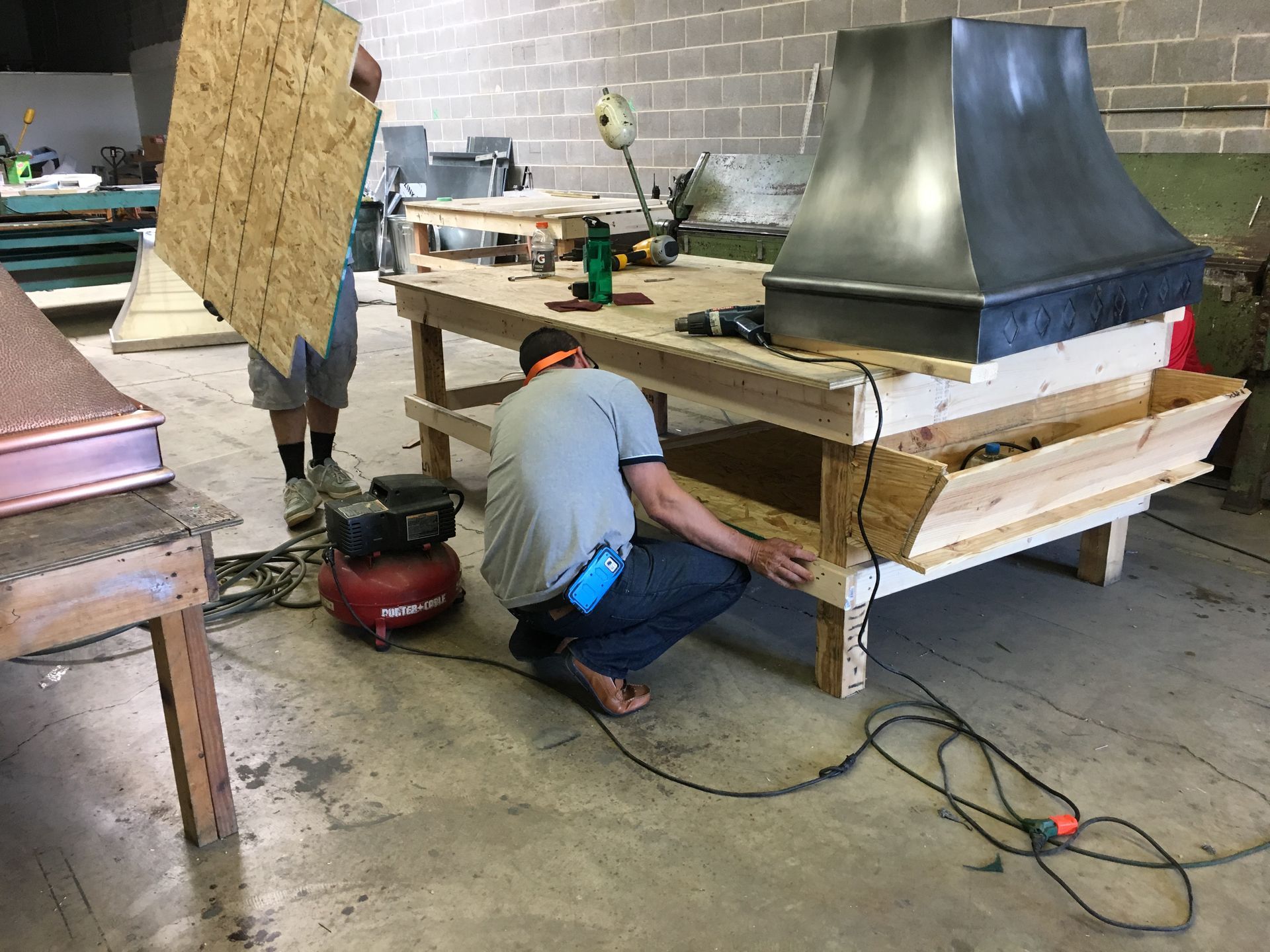 Worker crouches beside a large metal structure in a workshop, using tools on a wooden platform.