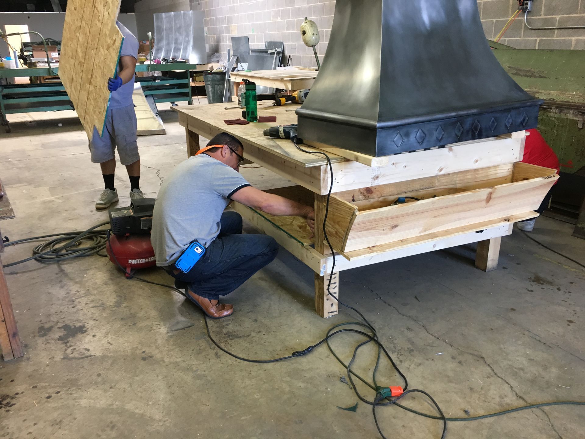 Workers assembling a large metal chimney hood on a wooden base in a workshop