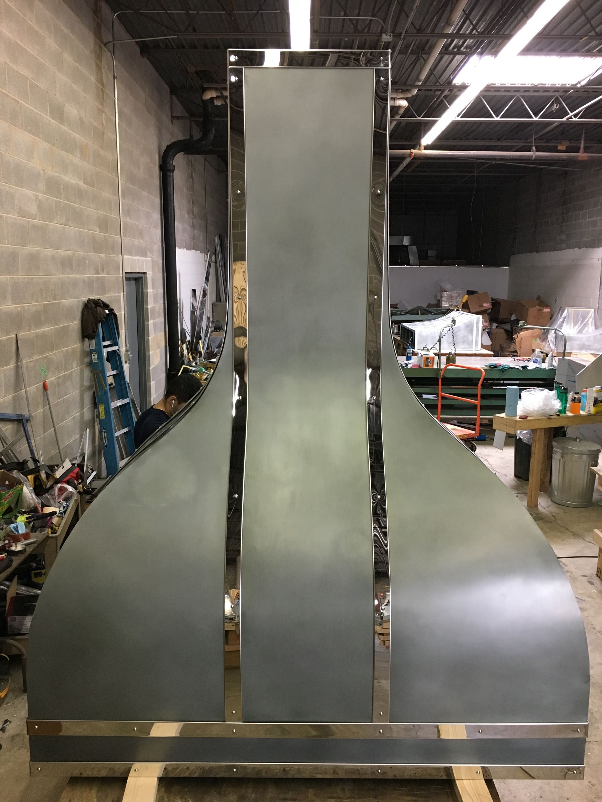 Large metal industrial hood or duct in a warehouse workshop, with shelves and tools in the background