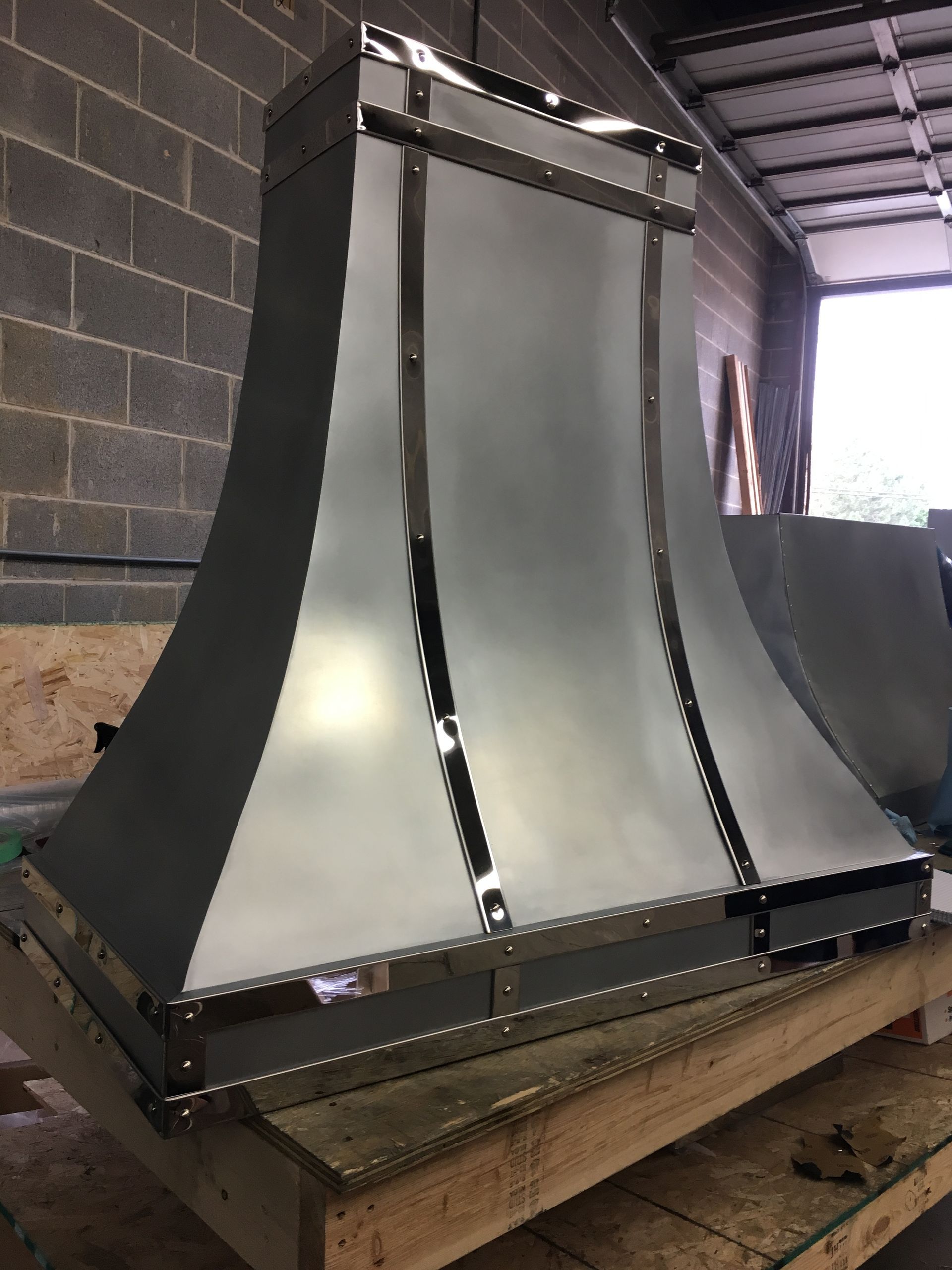 Large metal industrial duct or hood on a wooden pallet in a workshop, against a brick wall.