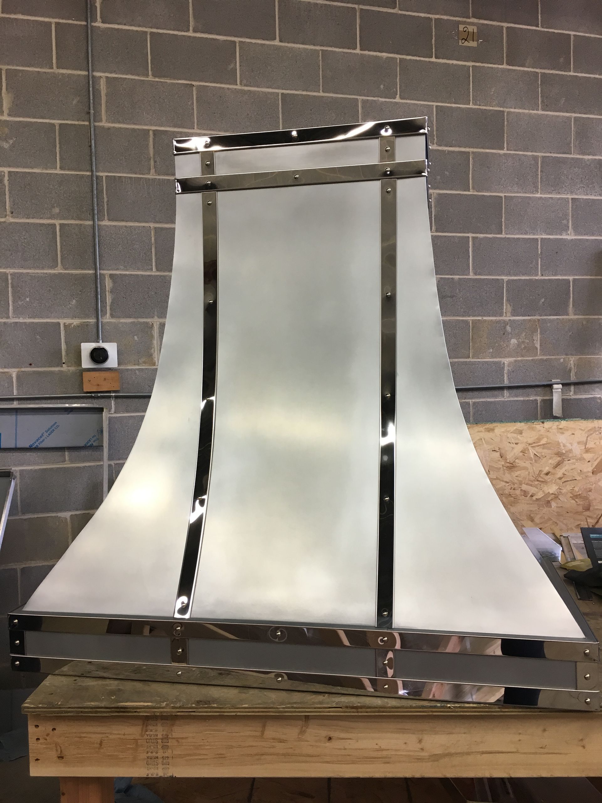 Metal prototype panel with curved silver sides on a wooden pallet against a cinderblock wall