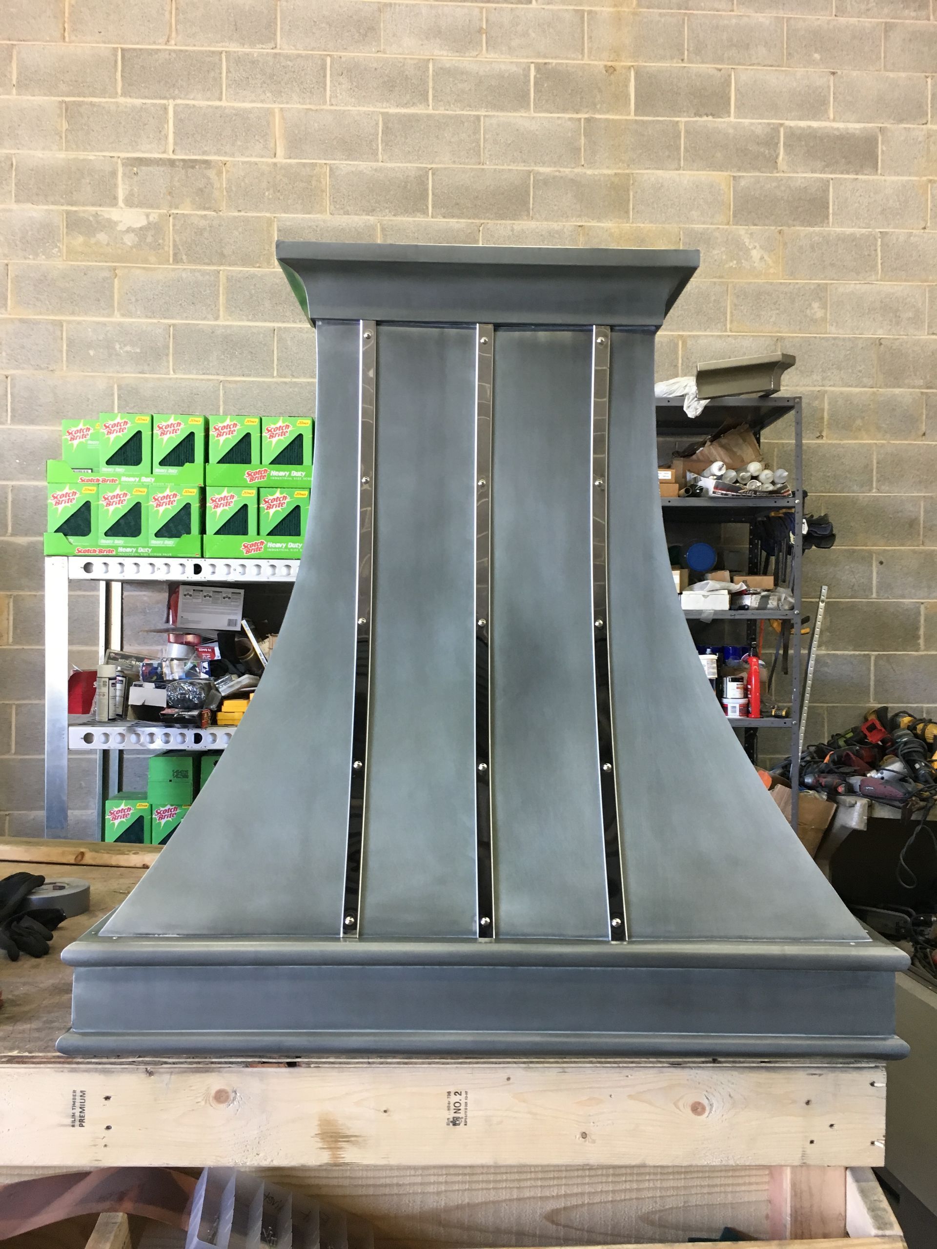 Gray metal range hood in a workshop, standing on a wooden pallet against a brick wall.
