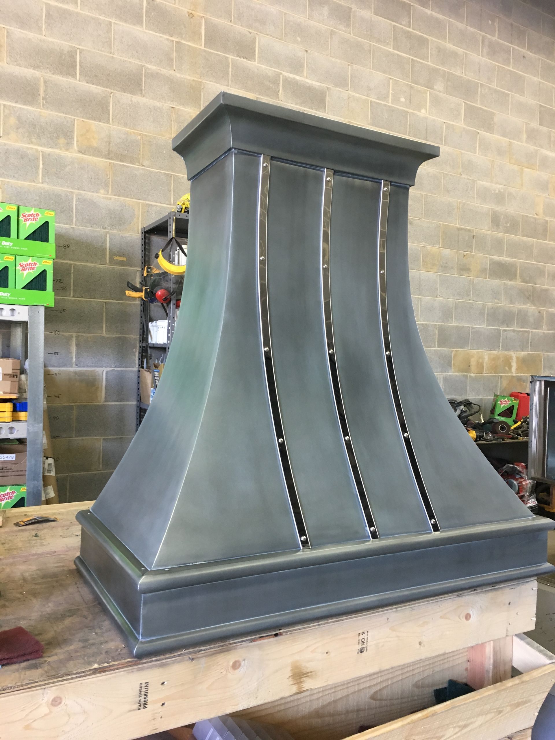 Gray metal stove hood with decorative trim, sitting on a workbench in a workshop.