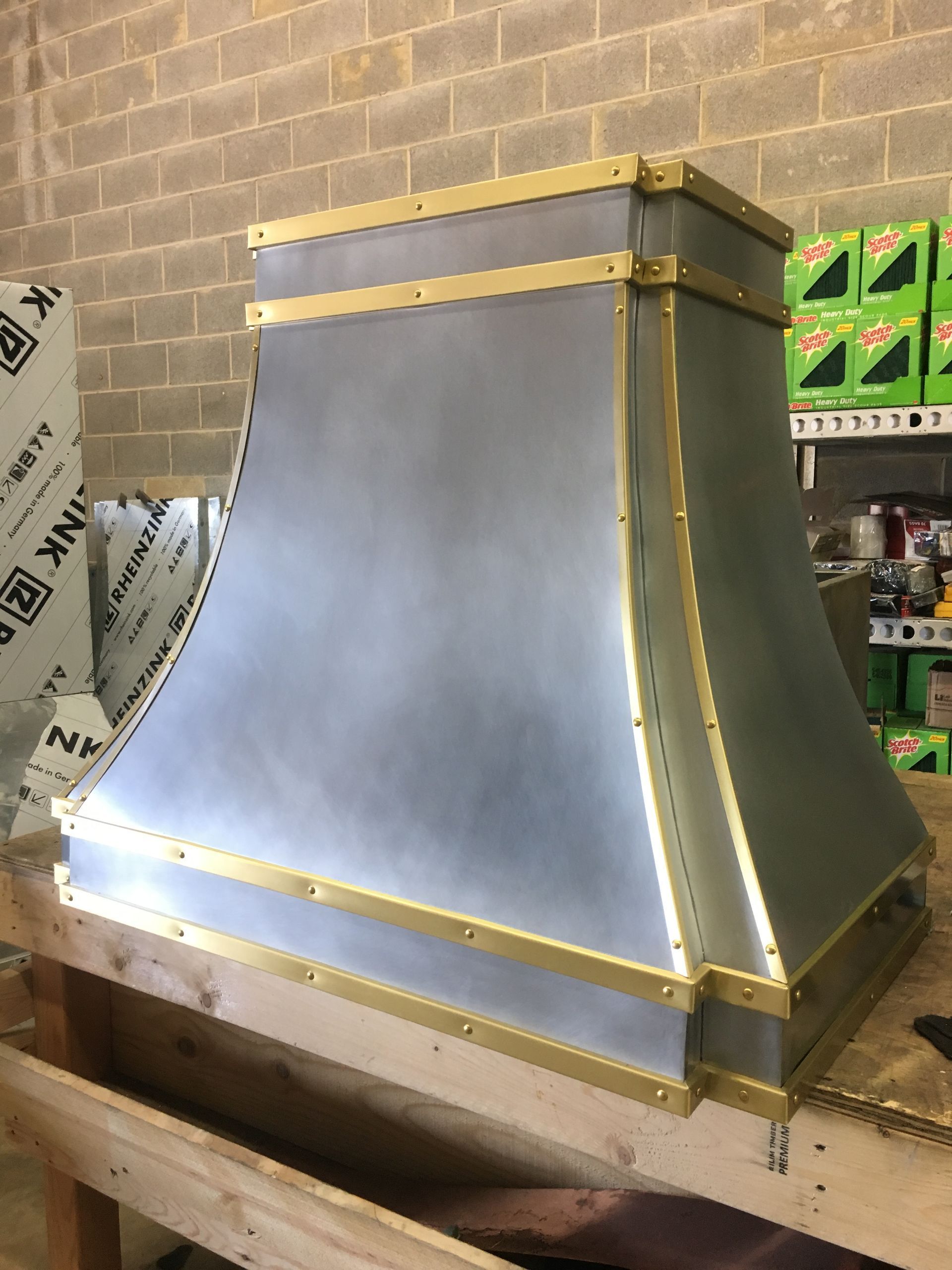 Large gray metal hood or chimney cap with brass trim, sitting on a workbench in a workshop.