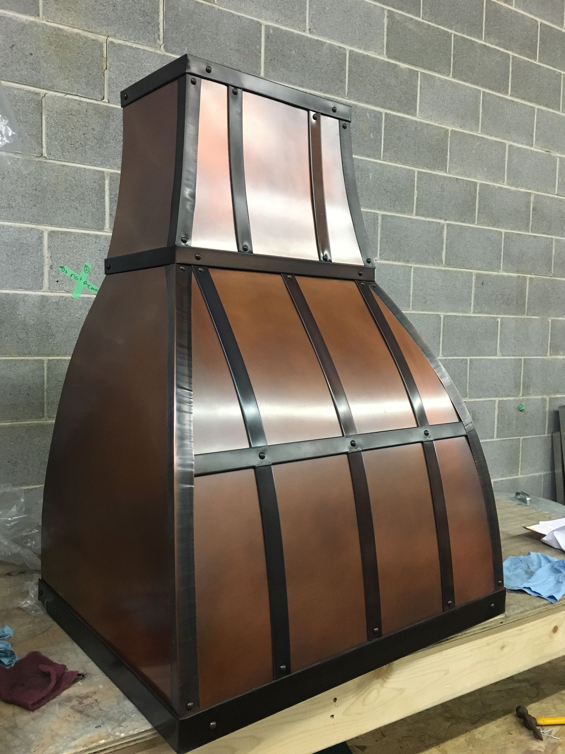 Copper-and-black metal hood or chimney cover standing on a workbench against a cinderblock wall.