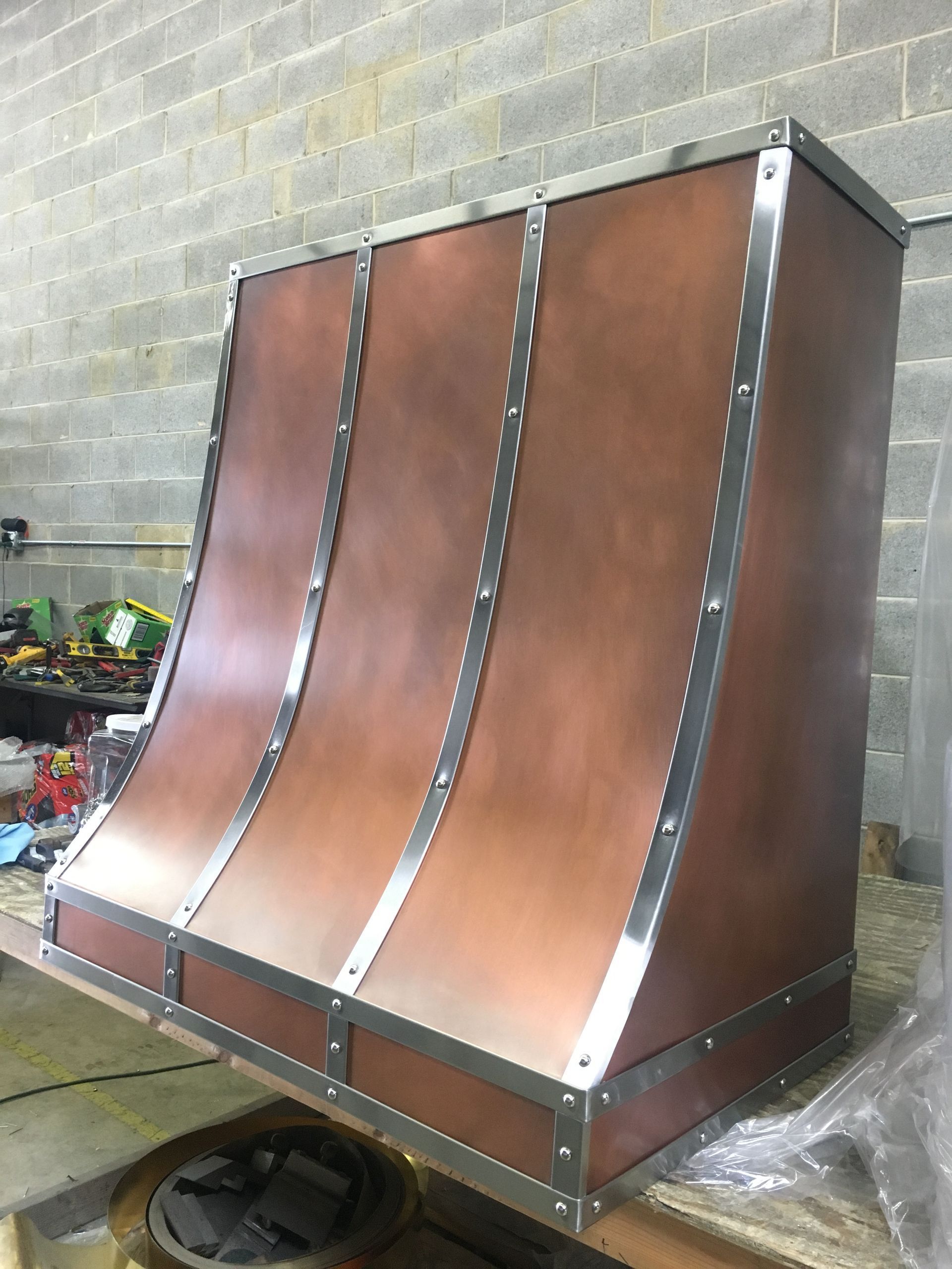 Copper-colored industrial metal hood with three curved panels, leaning against a concrete wall in a workshop.
