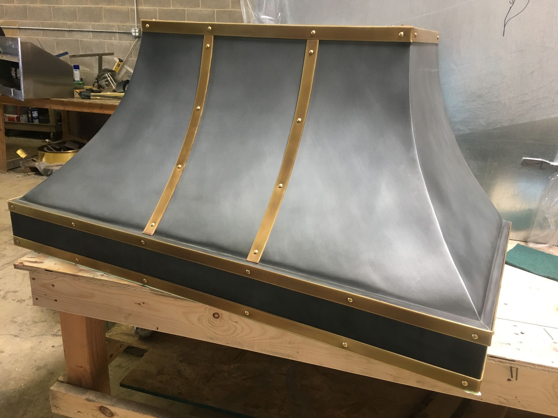 Large gray and gold metal hood or chimney cap on a wooden workbench in a workshop