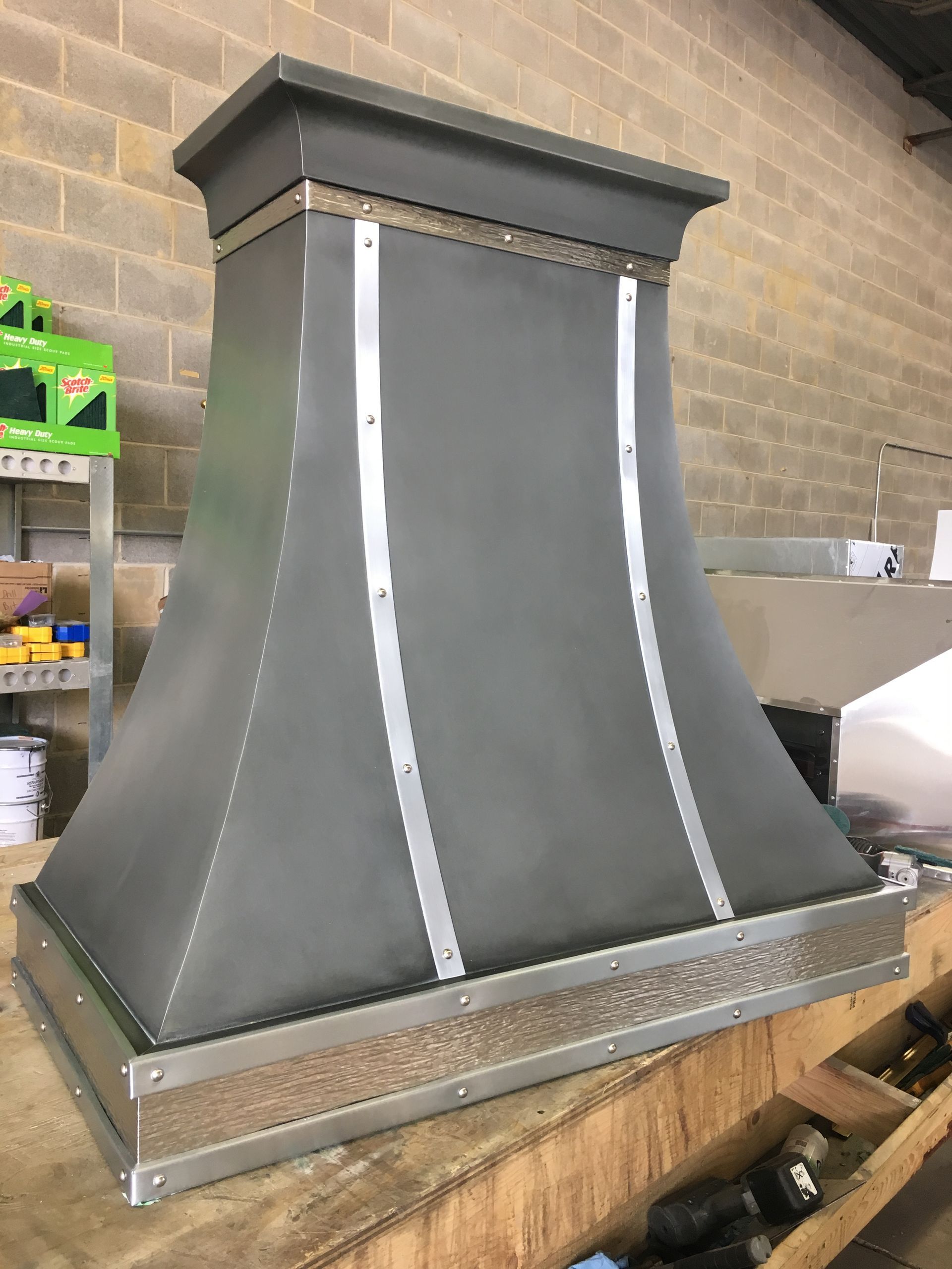 Gray metal range hood with a flared base and rectangular top, sitting in a workshop against a brick wall