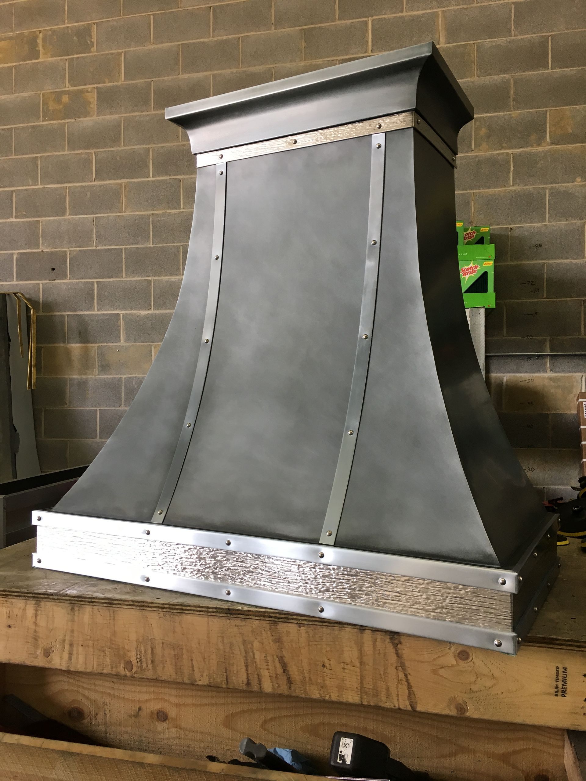 Metal range hood on a wooden workbench in a workshop, with unfinished sheet metal and brick wall background