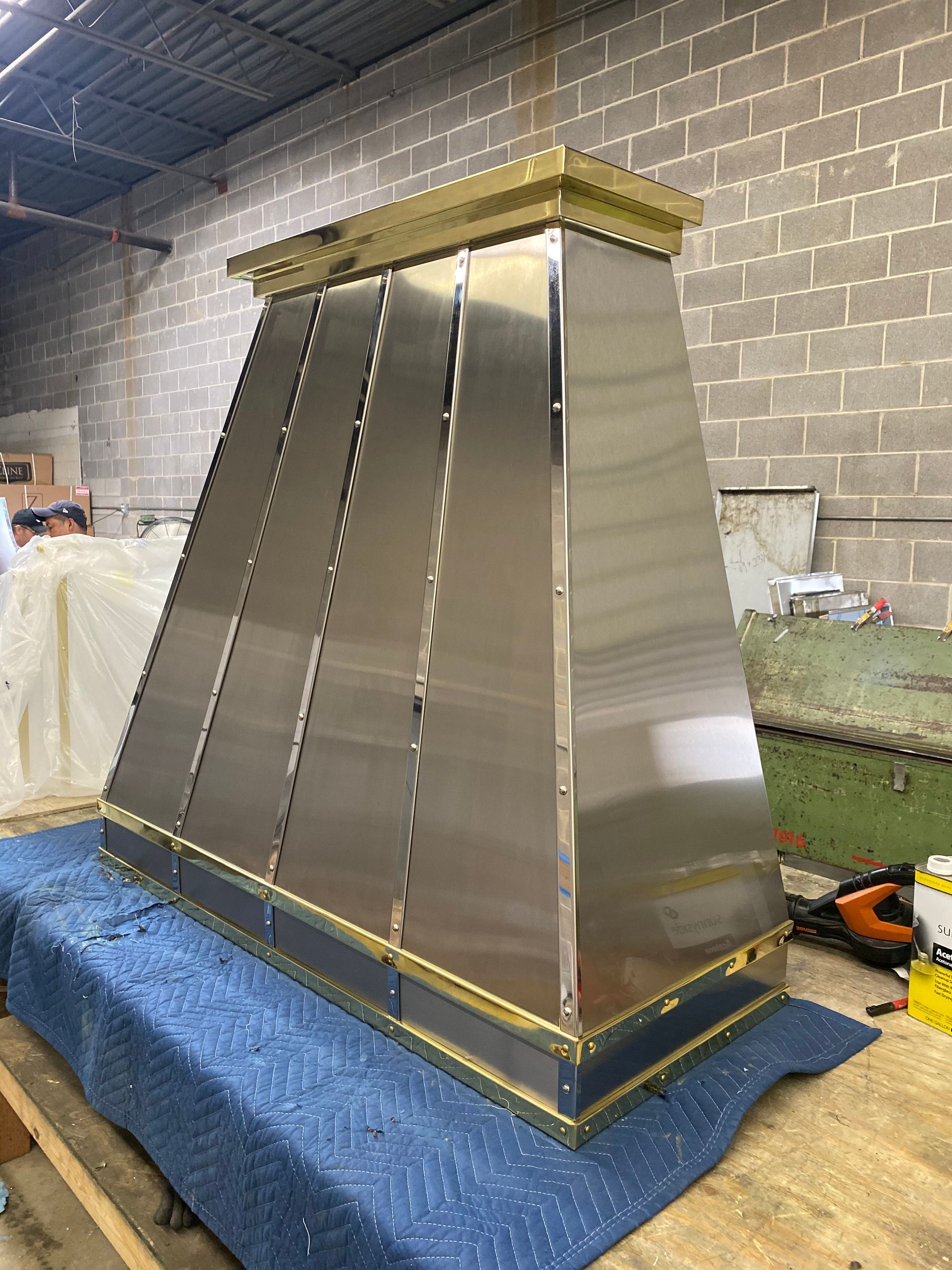 Large stainless steel chimney hood with gold trim sitting on a workshop floor.