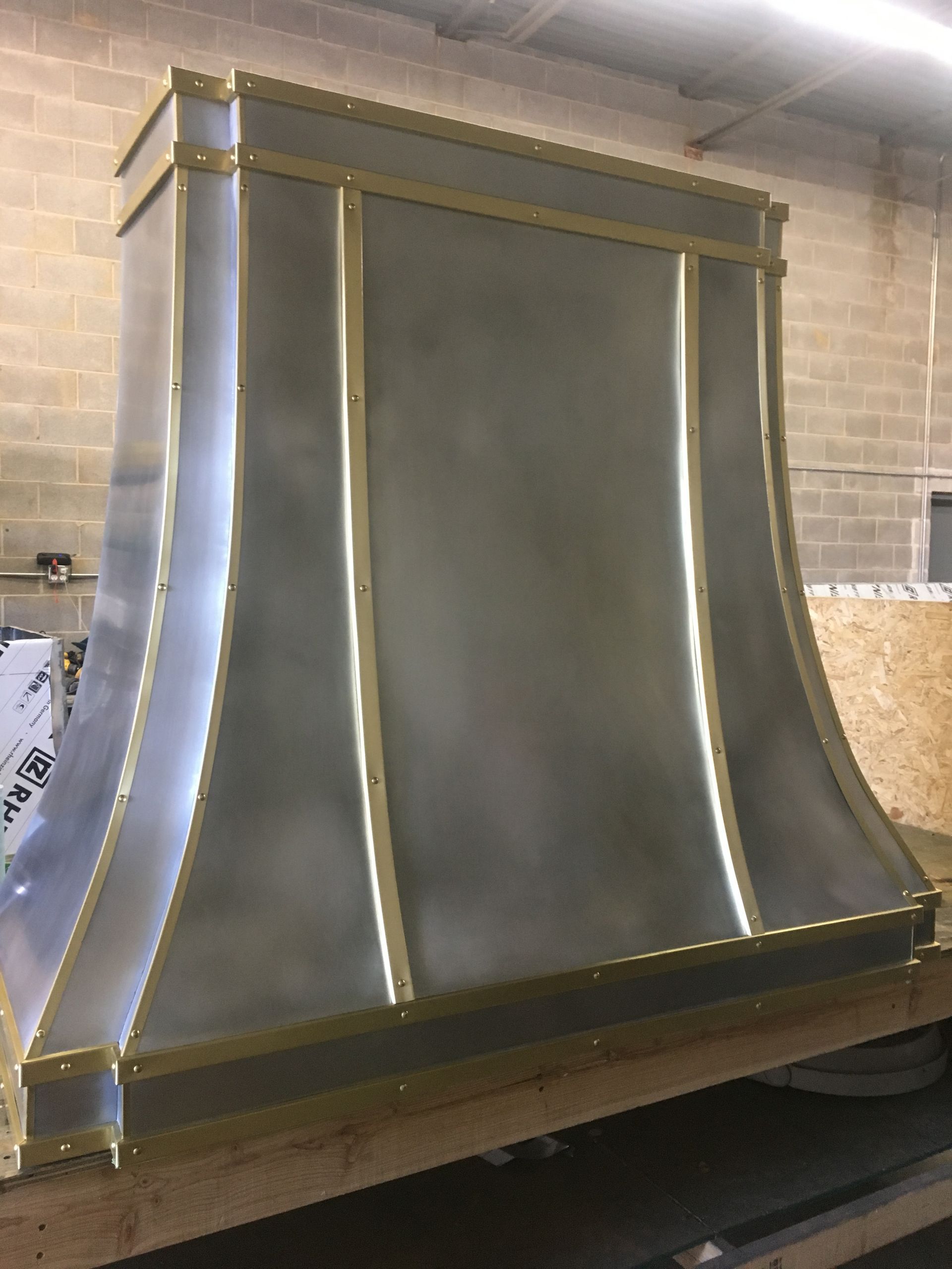 Silver metal fireplace hood with gold trim against a brick wall, sitting on a wooden base.