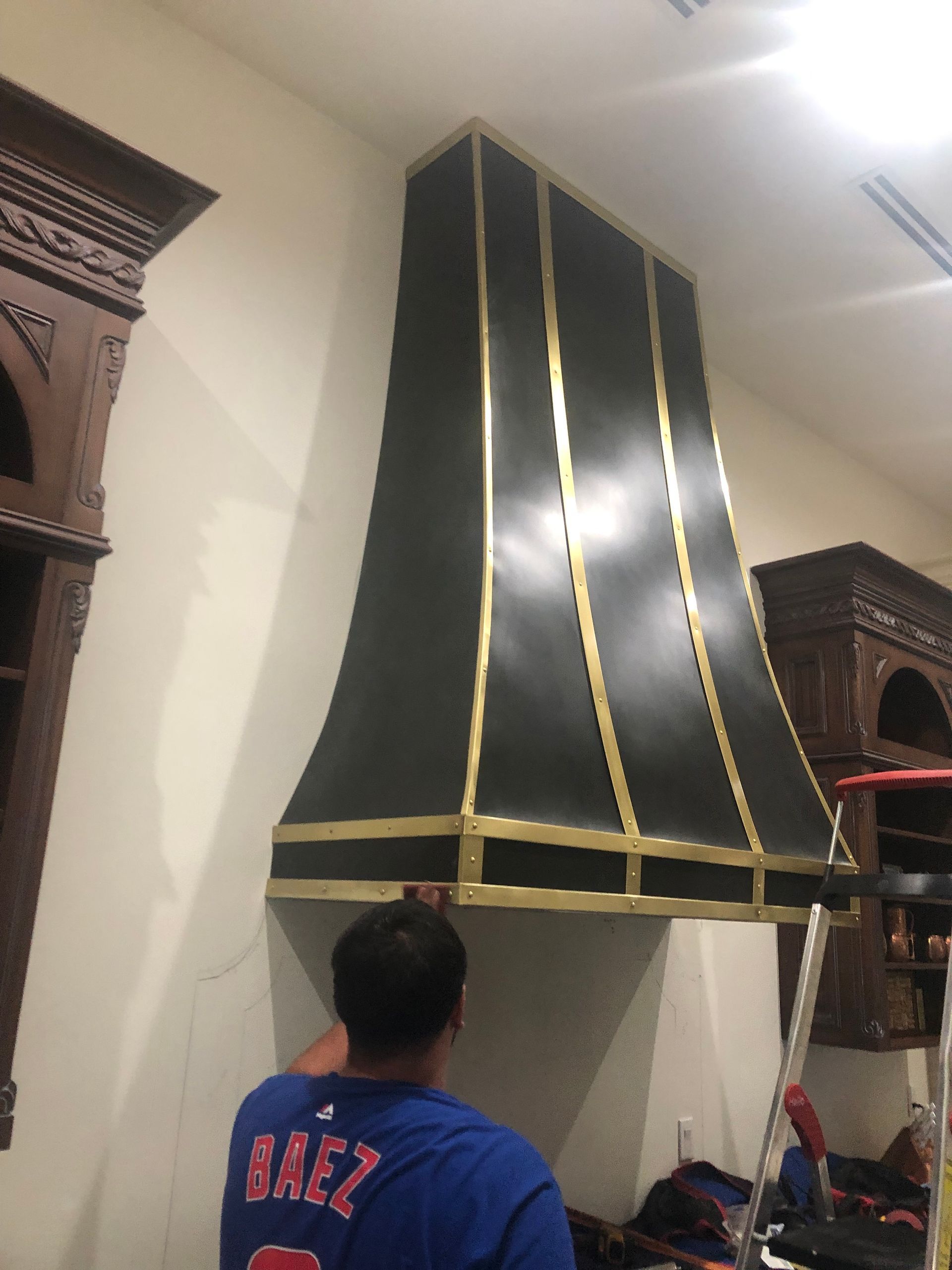 Person standing in a room, looking up at a large black chimney hood with gold trim.