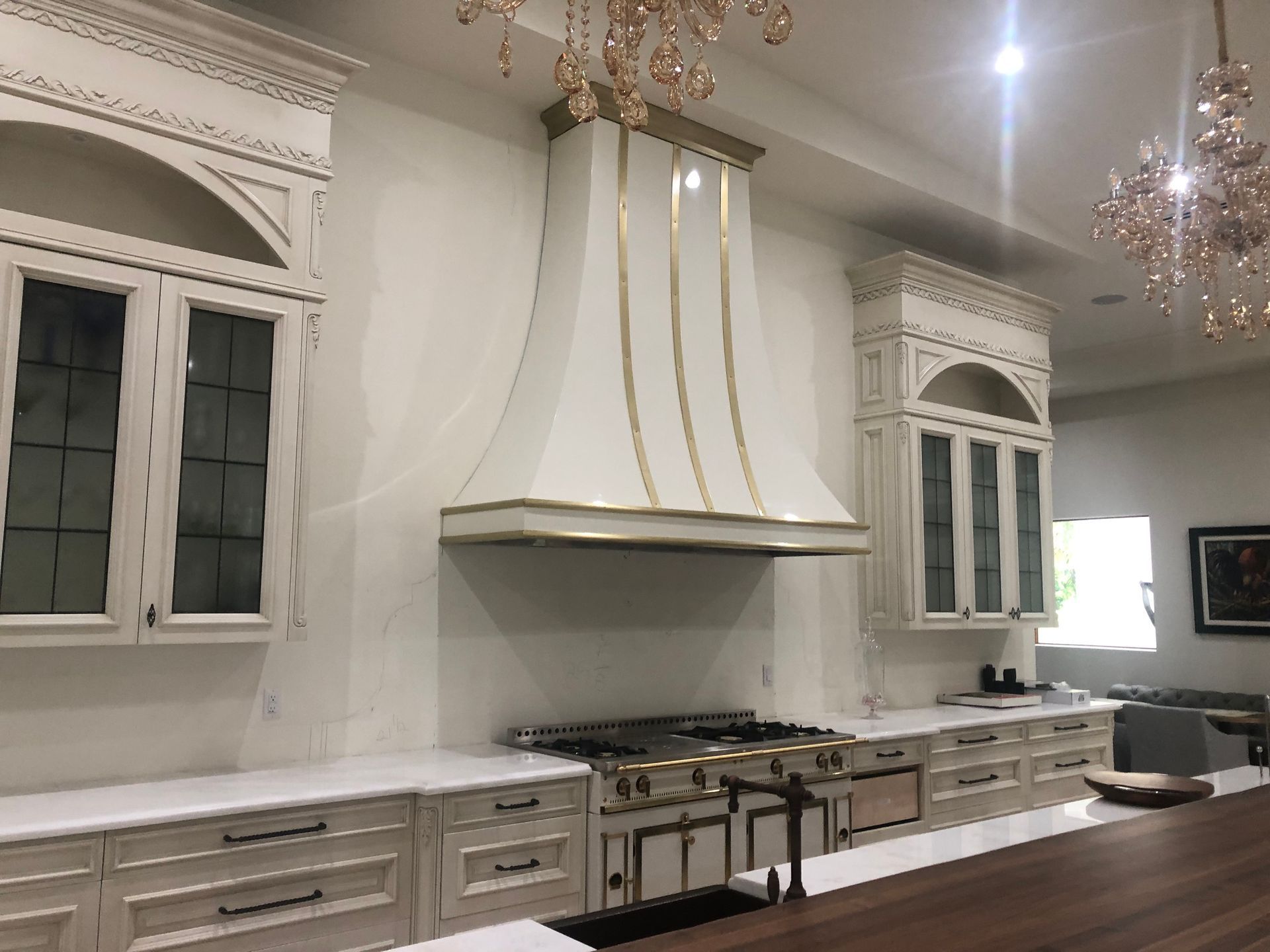 Elegant white kitchen with ornate cabinets, gold accents, large range hood, and chandelier lighting