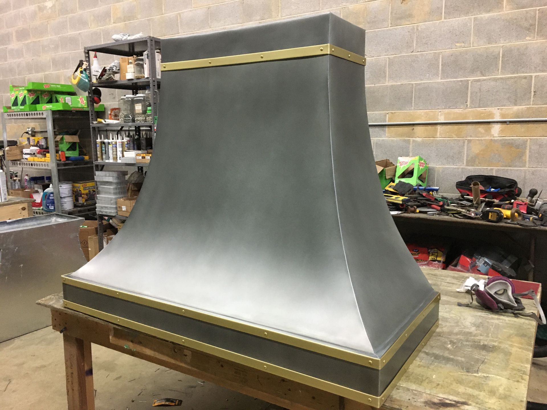 Large gray metal hood on a wooden workbench in a workshop, with gold trim and shelves in the background