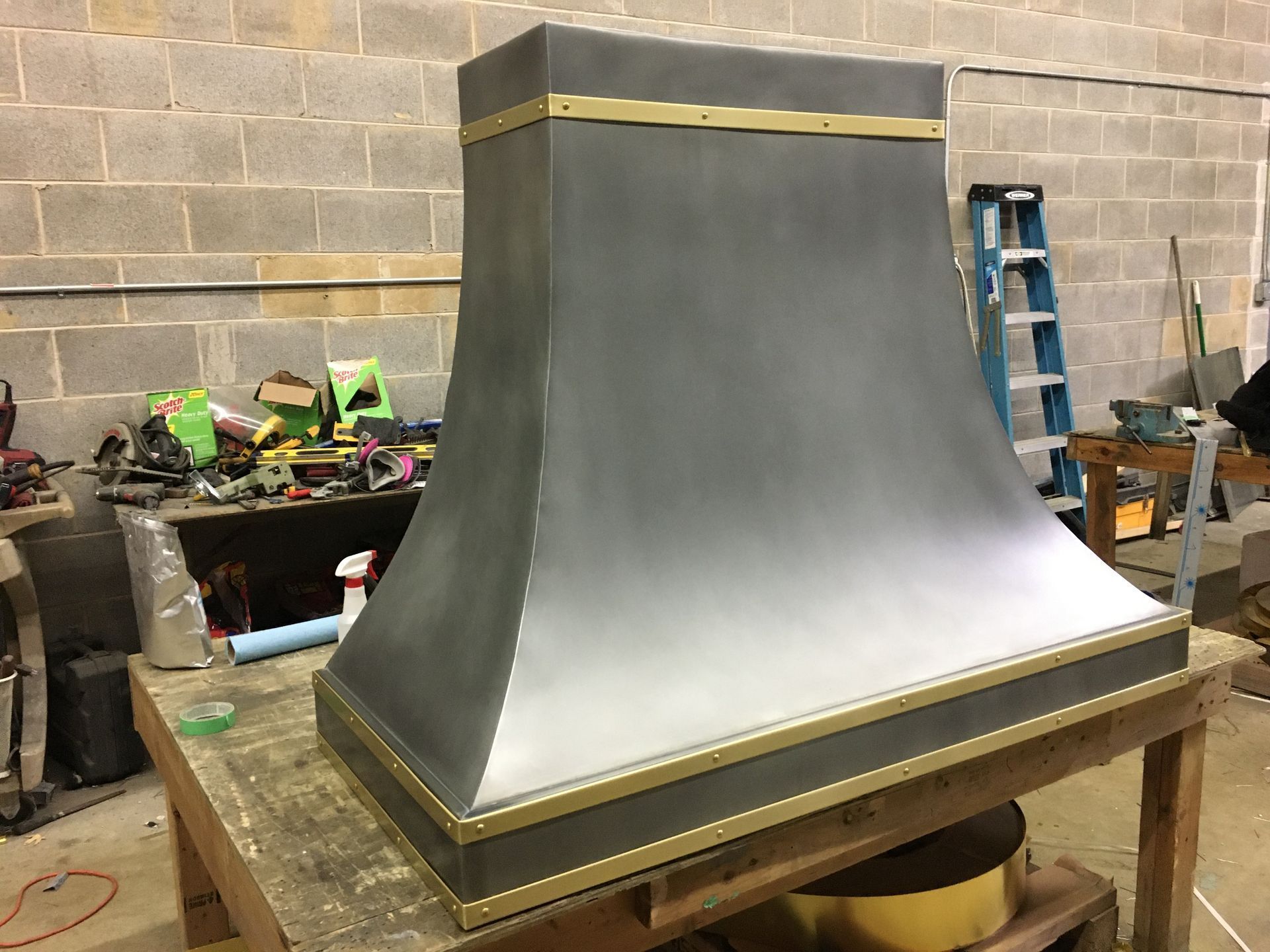 Large gray metal hood or chimney structure with gold trim on a workbench in a workshop