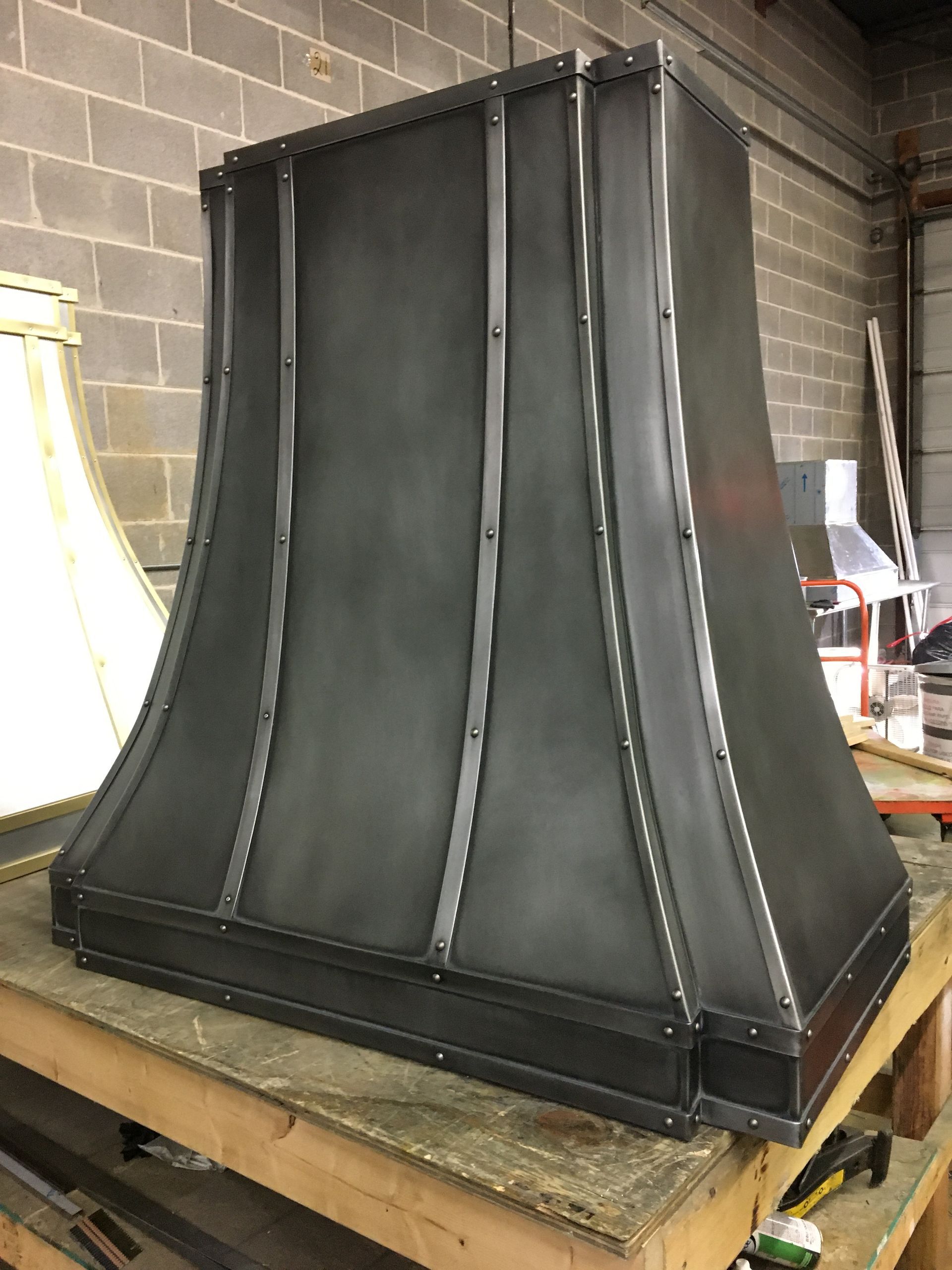 Large black metal hood or chimney shroud standing on a wooden pallet in a workshop.