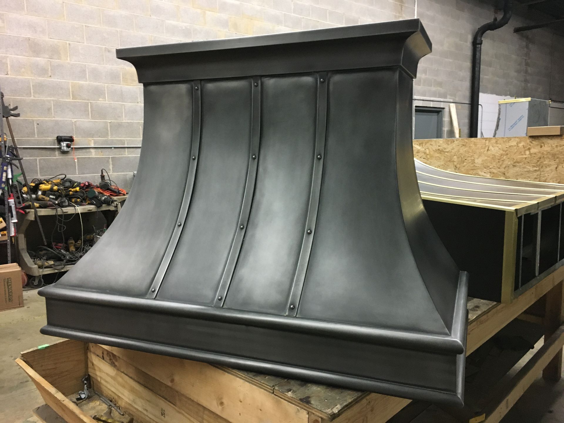 Dark gray metal range hood in a workshop, sitting on a wooden pallet with tools and materials nearby
