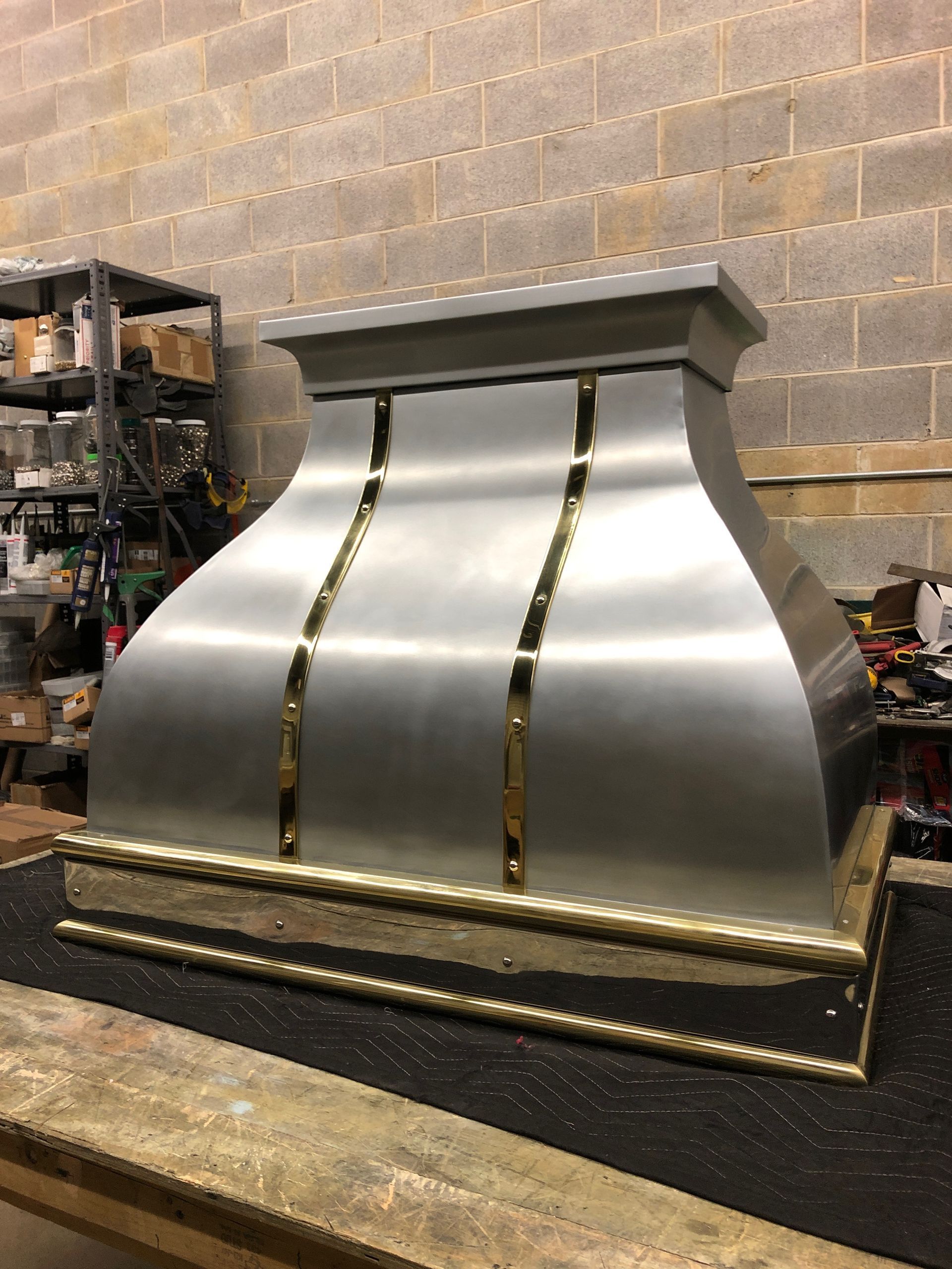 Large silver metal hood or vent cover on a workbench in a workshop, with brick wall and shelves behind it