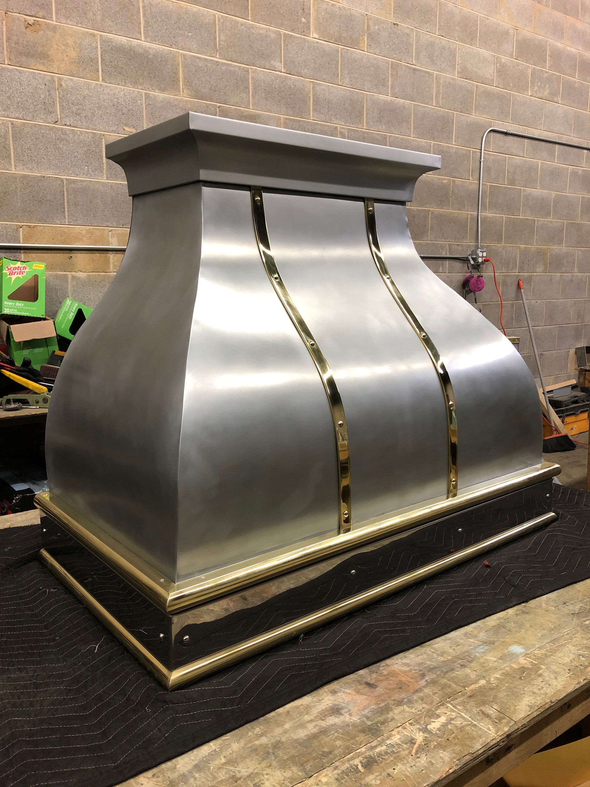 Large silver metal duct or chimney cap with gold trim in a workshop against a brick wall