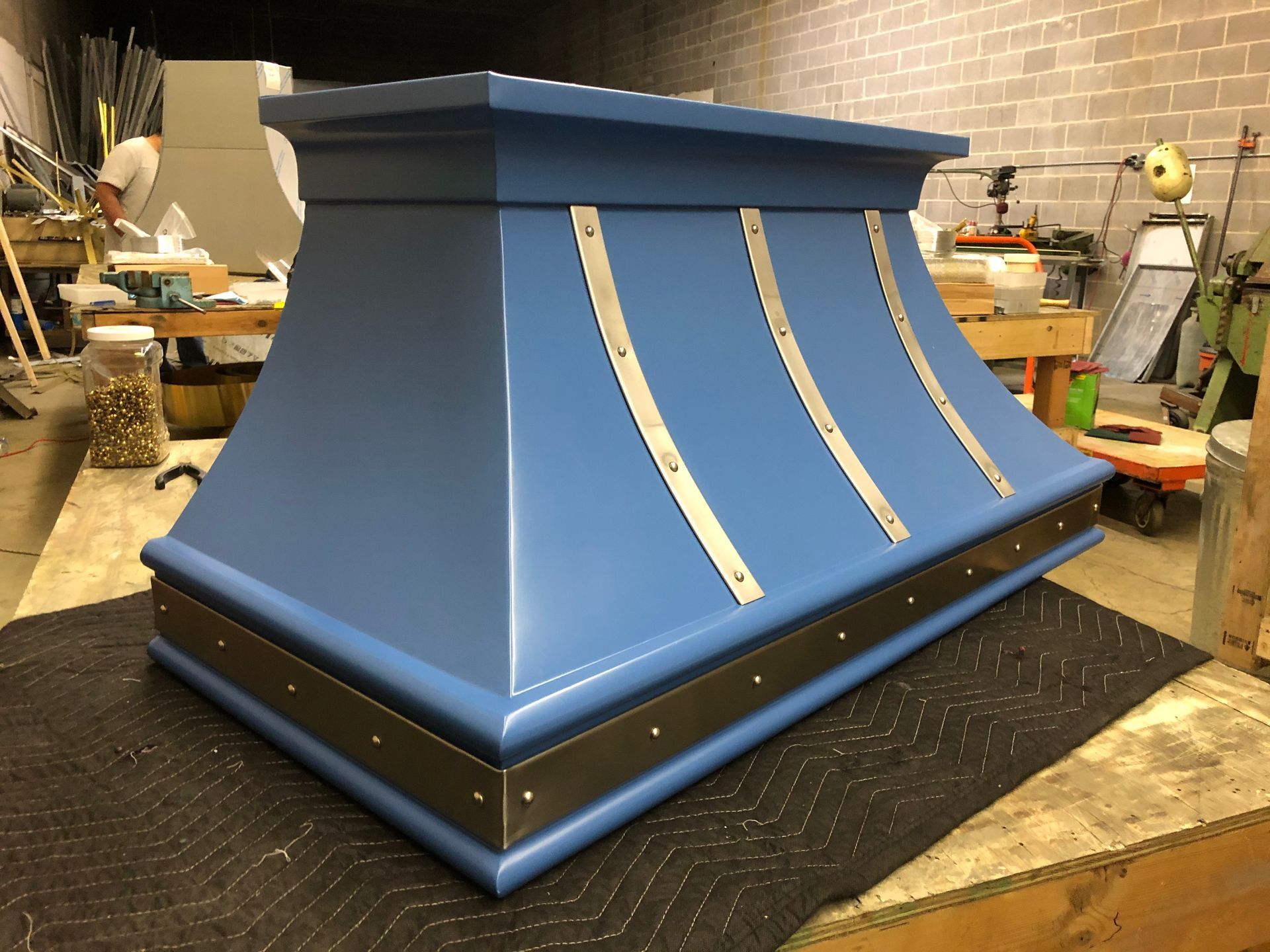 Blue metal hood or chimney cap with silver trim, shown on a workshop table.