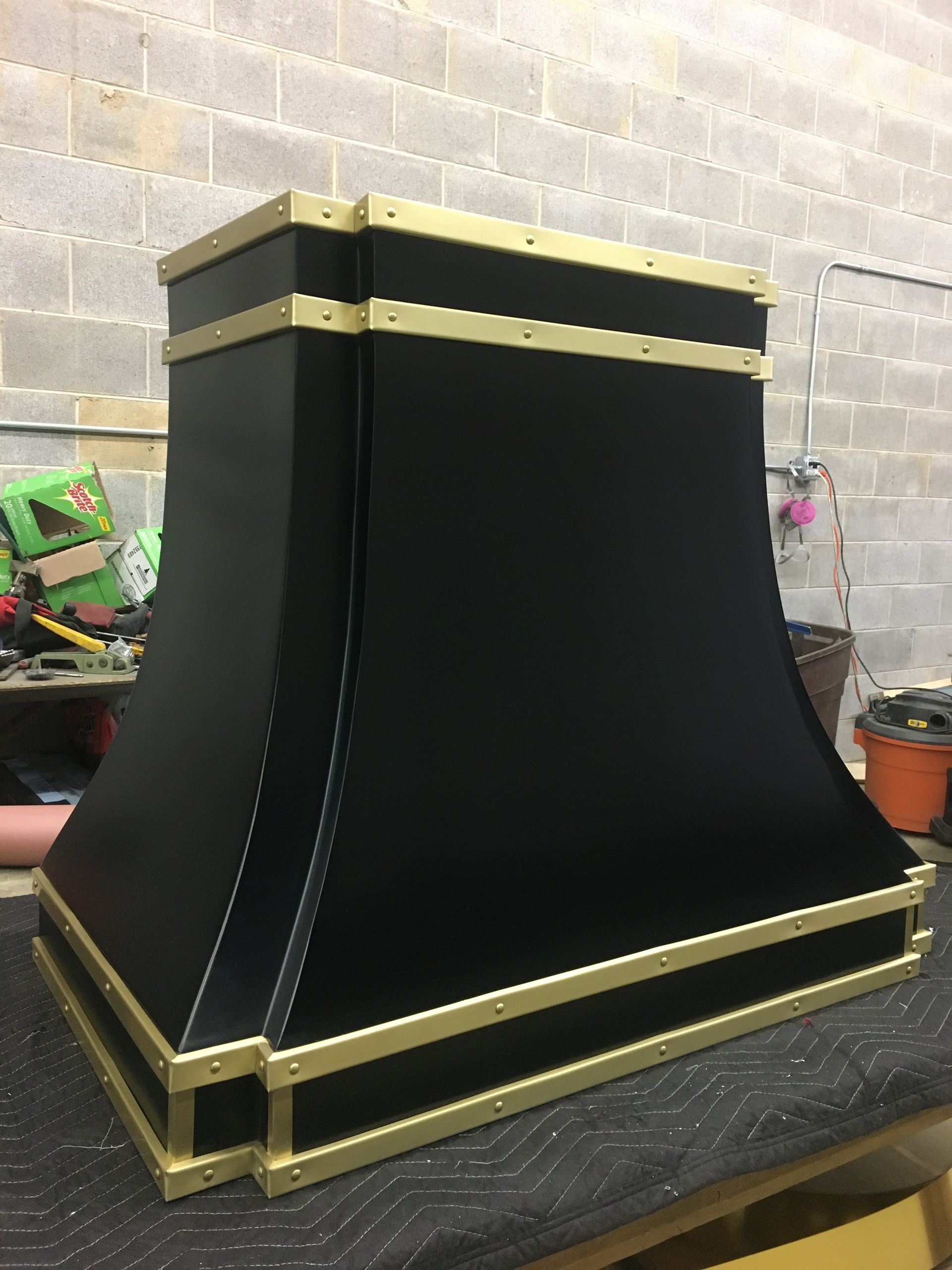 Black and gold metal fireplace hood on a workbench against a gray brick wall