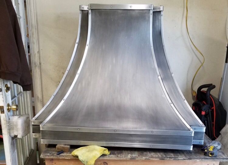 Stainless steel range hood under construction in a workshop, with curved sides and a square top.