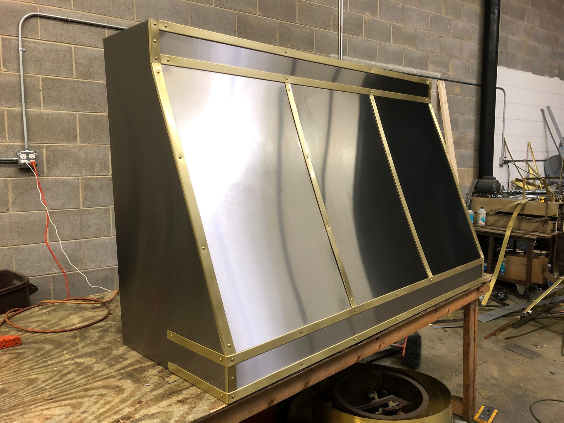 Large metal box or enclosure with slanted reflective panels in a workshop against a concrete wall.