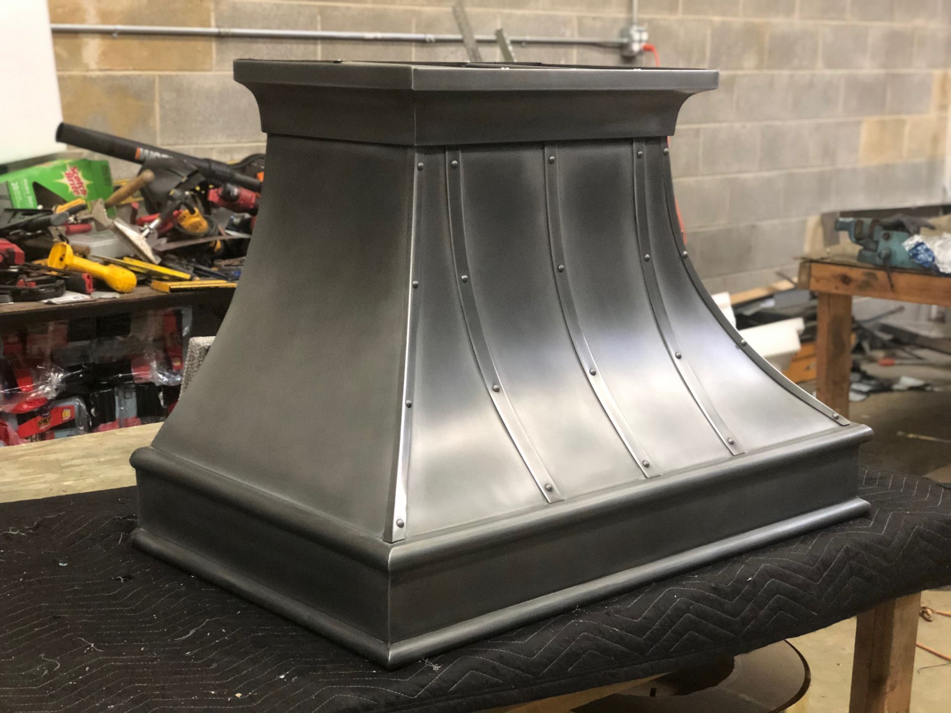 Large black metal vent hood in a workshop, with a flared base and riveted panels.