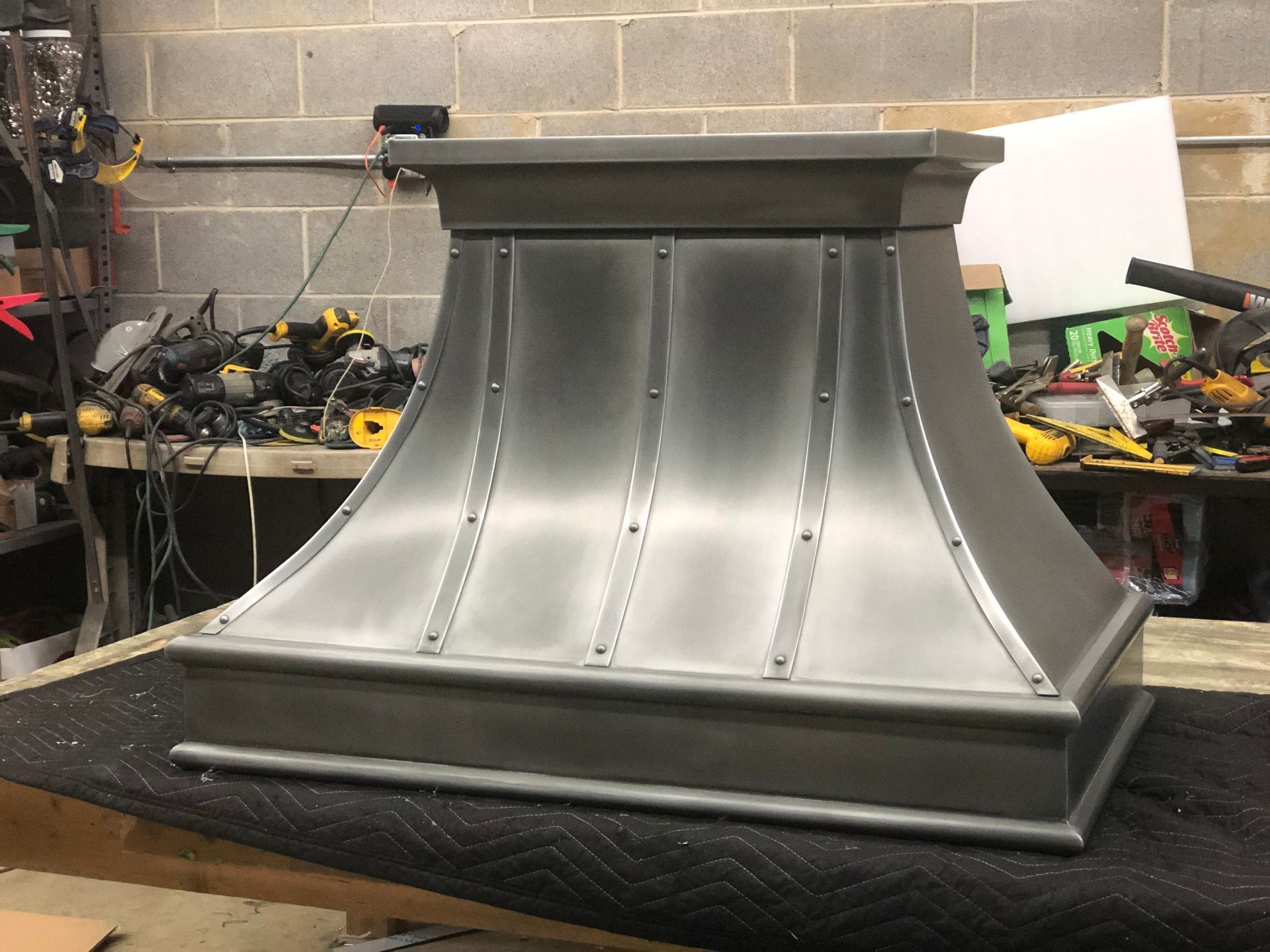 Gray metal chimney hood or roof vent on a workbench in a workshop, with tools in the background