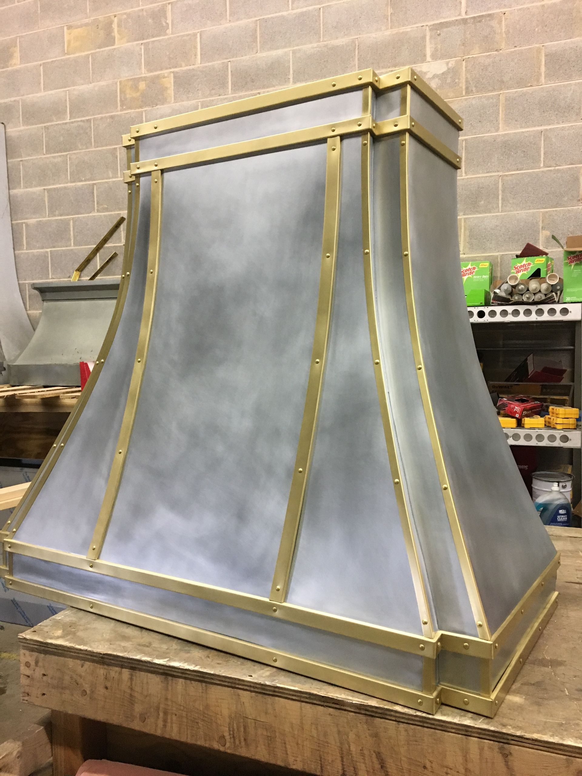 Metal chimney hood with brass trim on a wooden workbench in a workshop
