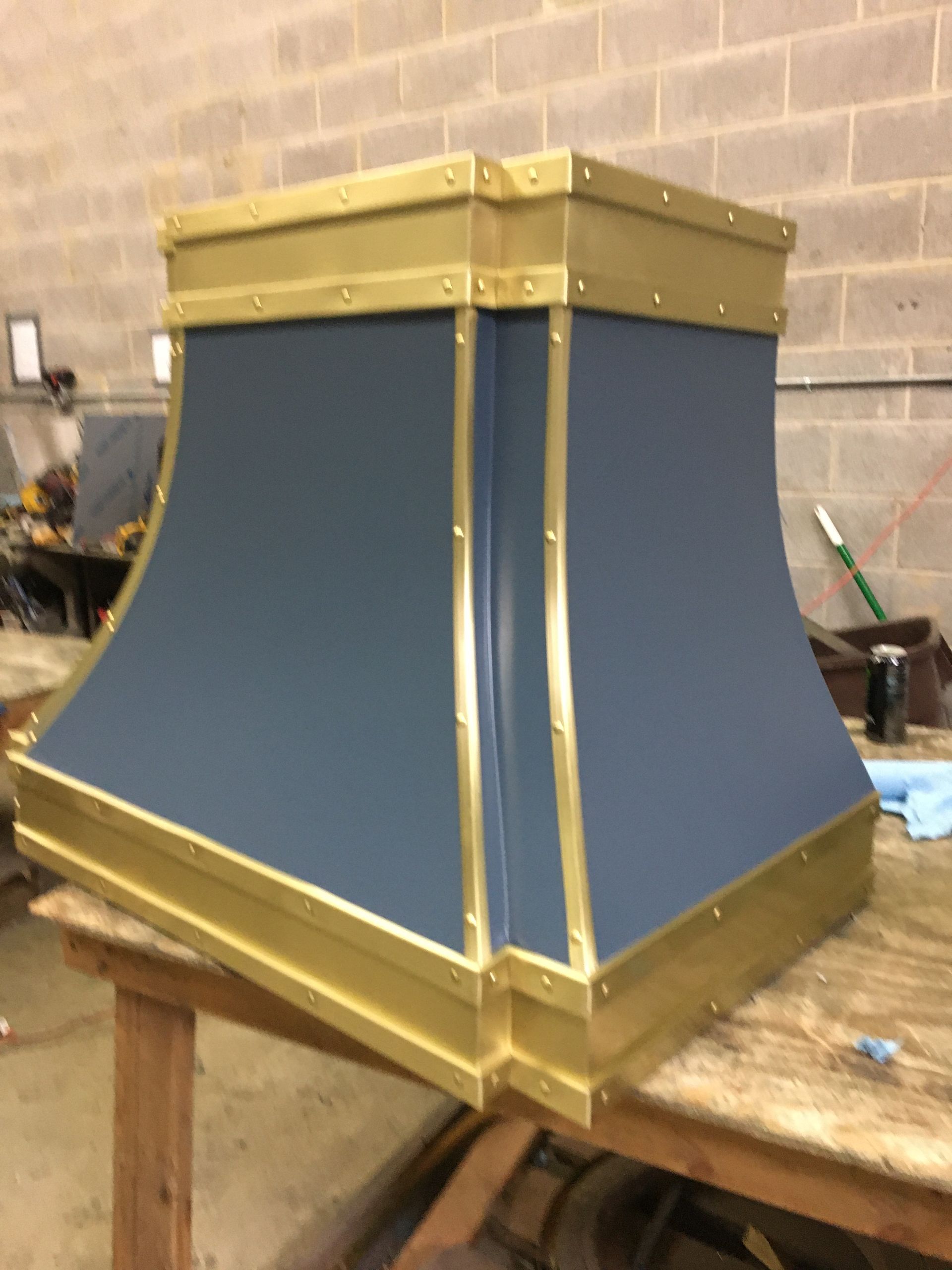 Blue and gold industrial metal enclosure on a workshop bench, with a tapered shape and riveted trim