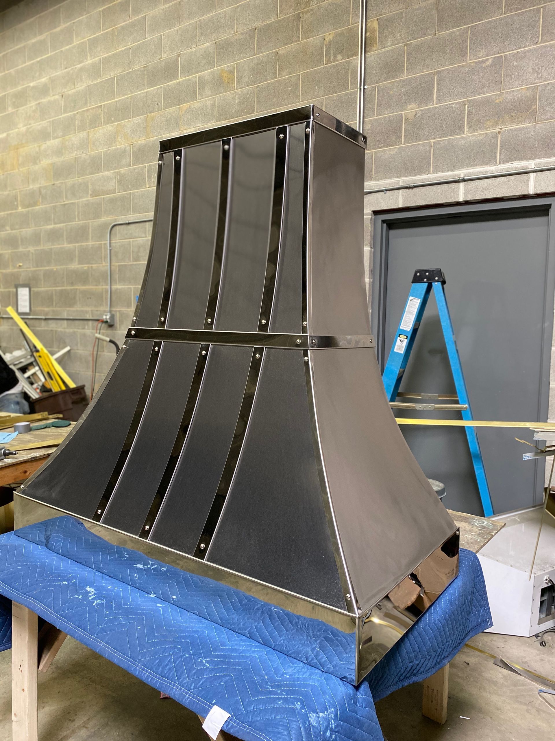 Large black metal structure on a blue pallet in a workshop, with a ladder and cinderblock wall behind it