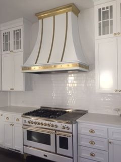 White kitchen with a stainless steel range, gold-trimmed vent hood, and white cabinetry