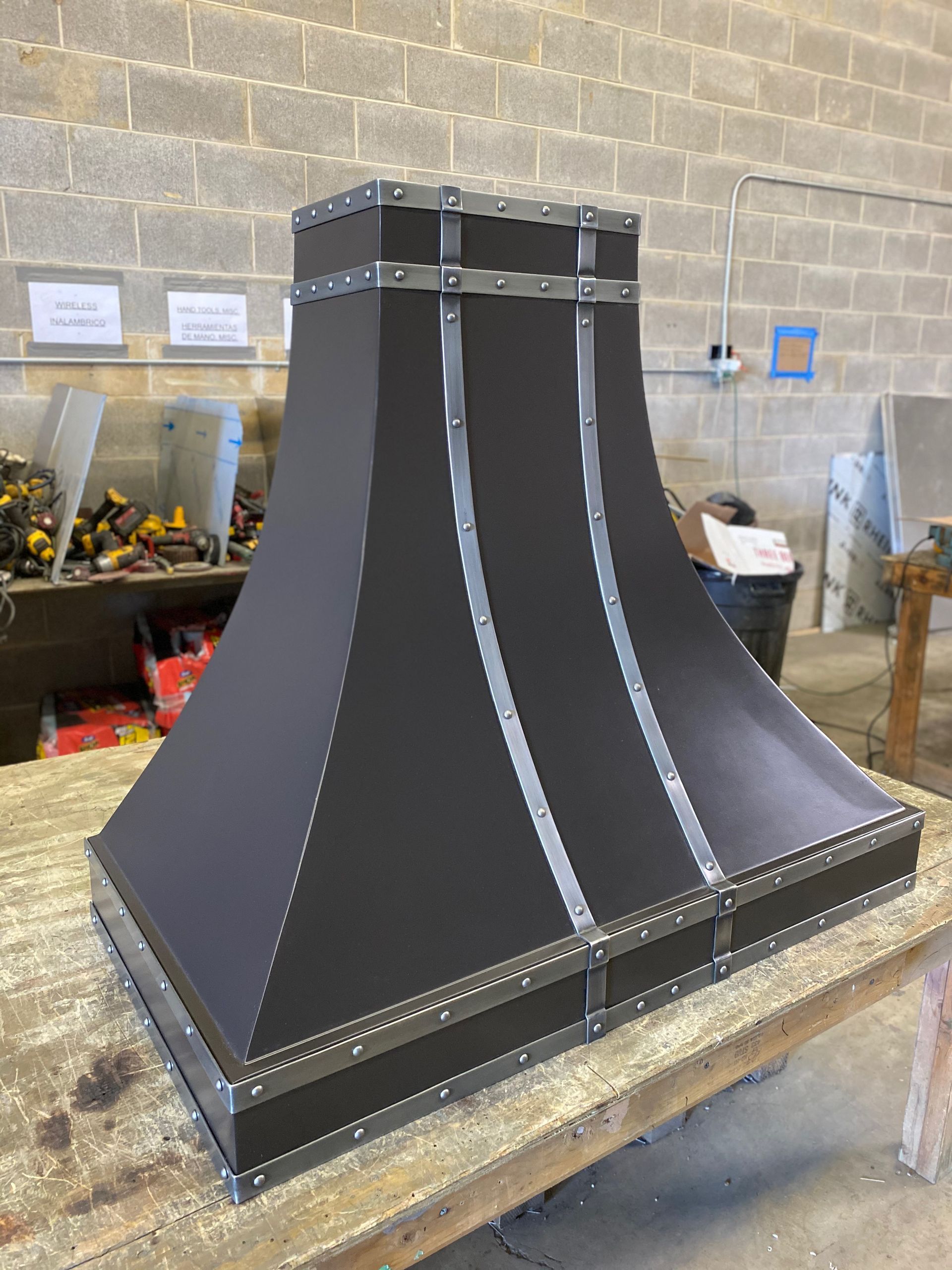 Large black metal chimney hood or vent structure in a workshop, with silver trim and riveted seams.
