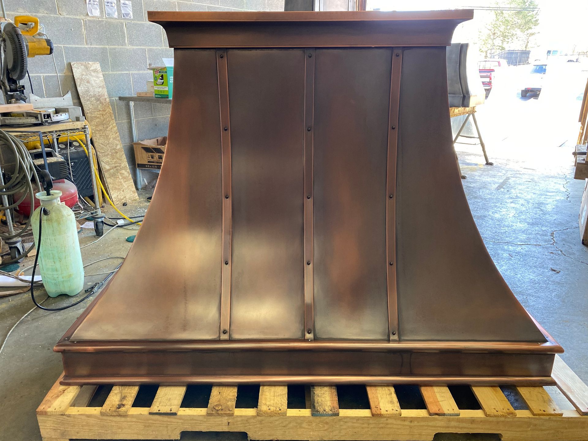 Large dark wood kitchen range hood on a pallet in a workshop, with a curved canopy and flat top.