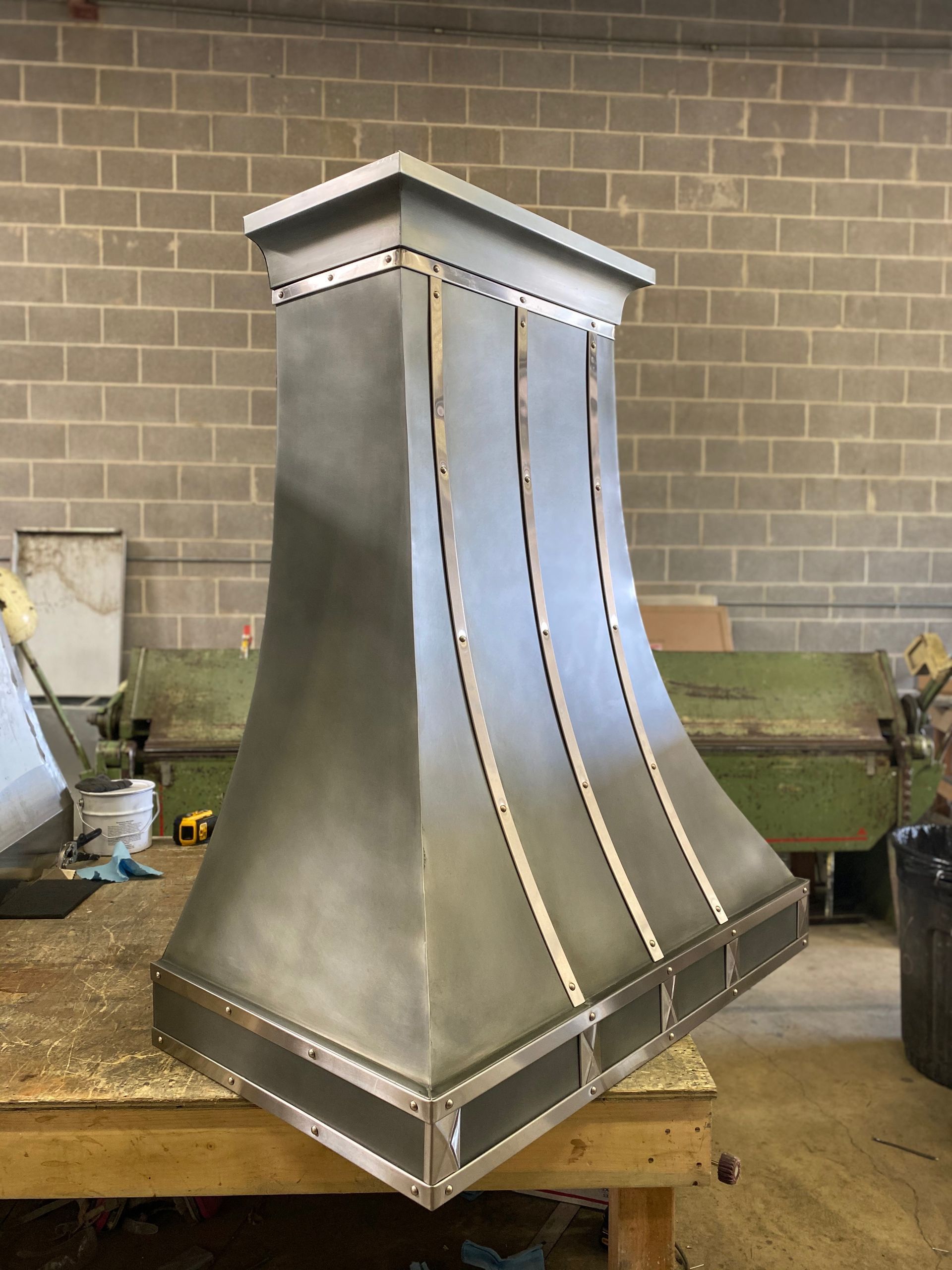 Stainless steel decorative hood with curved panels on a workshop table, against a brick wall.