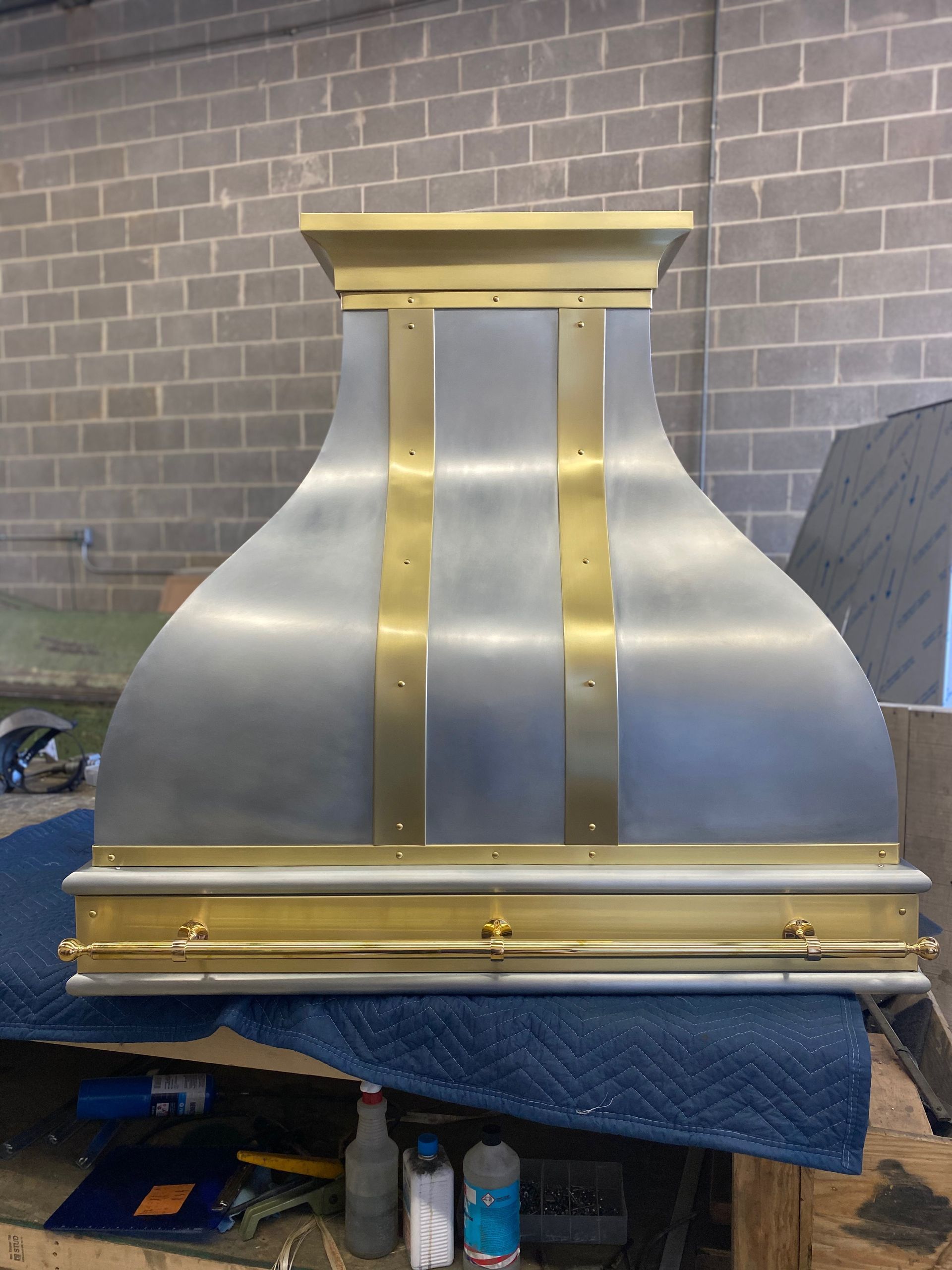 Brushed metal chimney hood with gold trim against a gray brick wall in a workshop