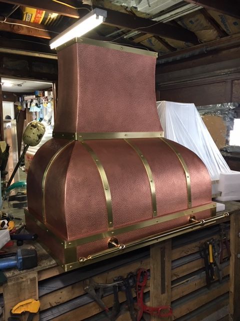 Copper-colored range hood in a workshop, with brass trim and a square chimney.