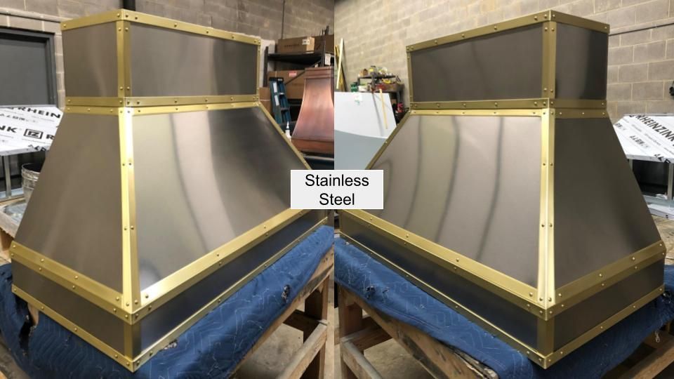 Two stainless steel vent hoods with gold trim on blue pallets in a workshop