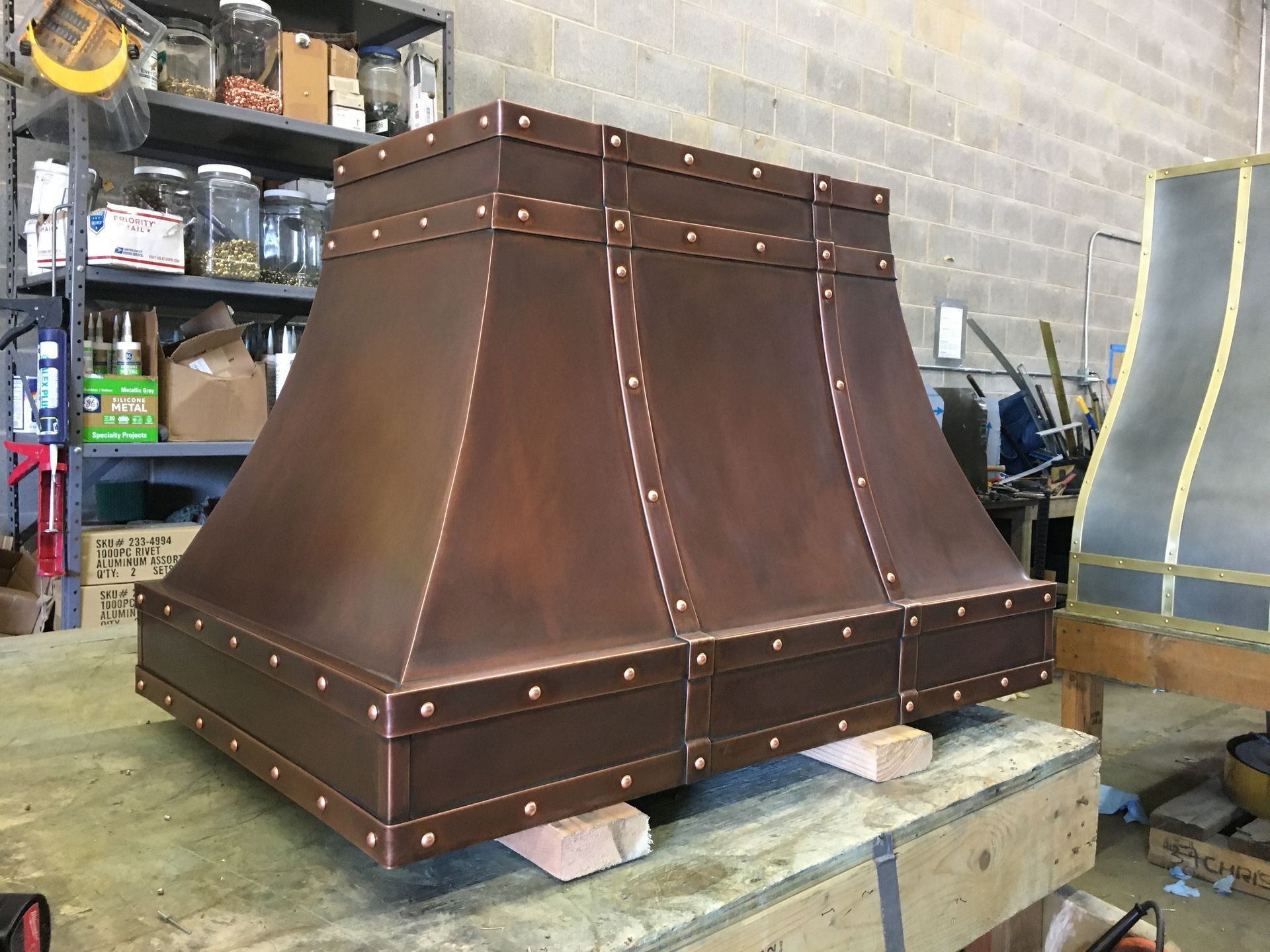 Copper-colored metal chimney hood on a pallet in a workshop, with riveted panels and shelves in the background