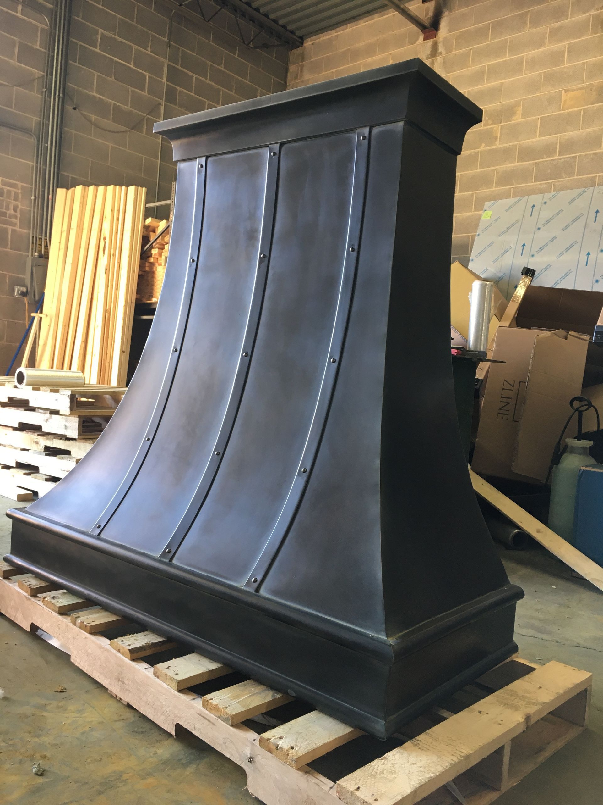 Large black decorative range hood or fireplace surround on a wooden pallet in a workshop.