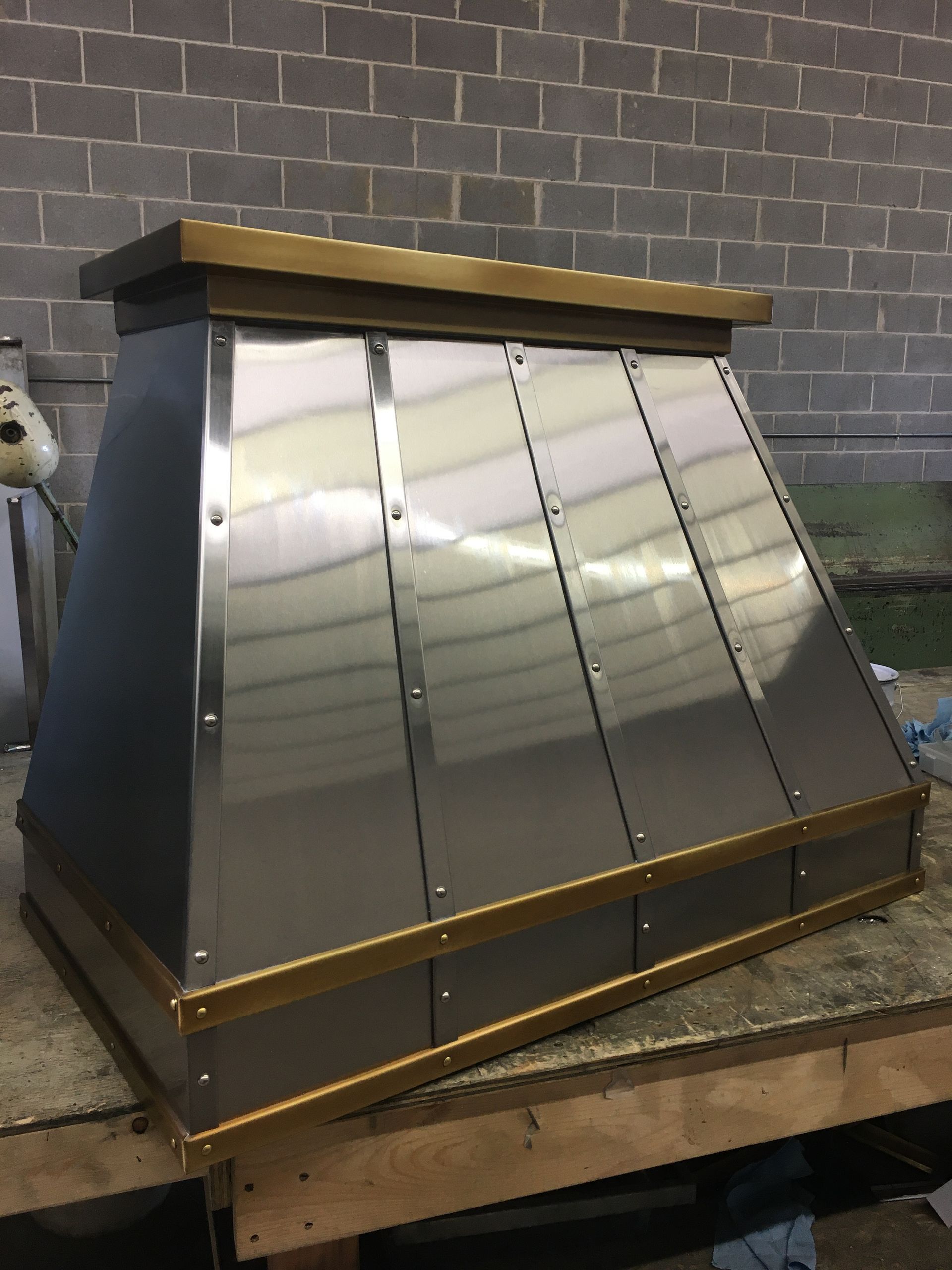 Metal chimney hood on a workbench, with shiny stainless panels and a dark top trim.