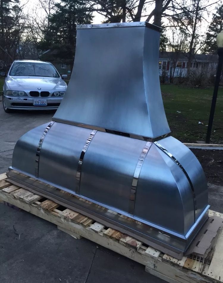 Large silver metal chimney hood on a wooden pallet outdoors, with a car and trees in the background.