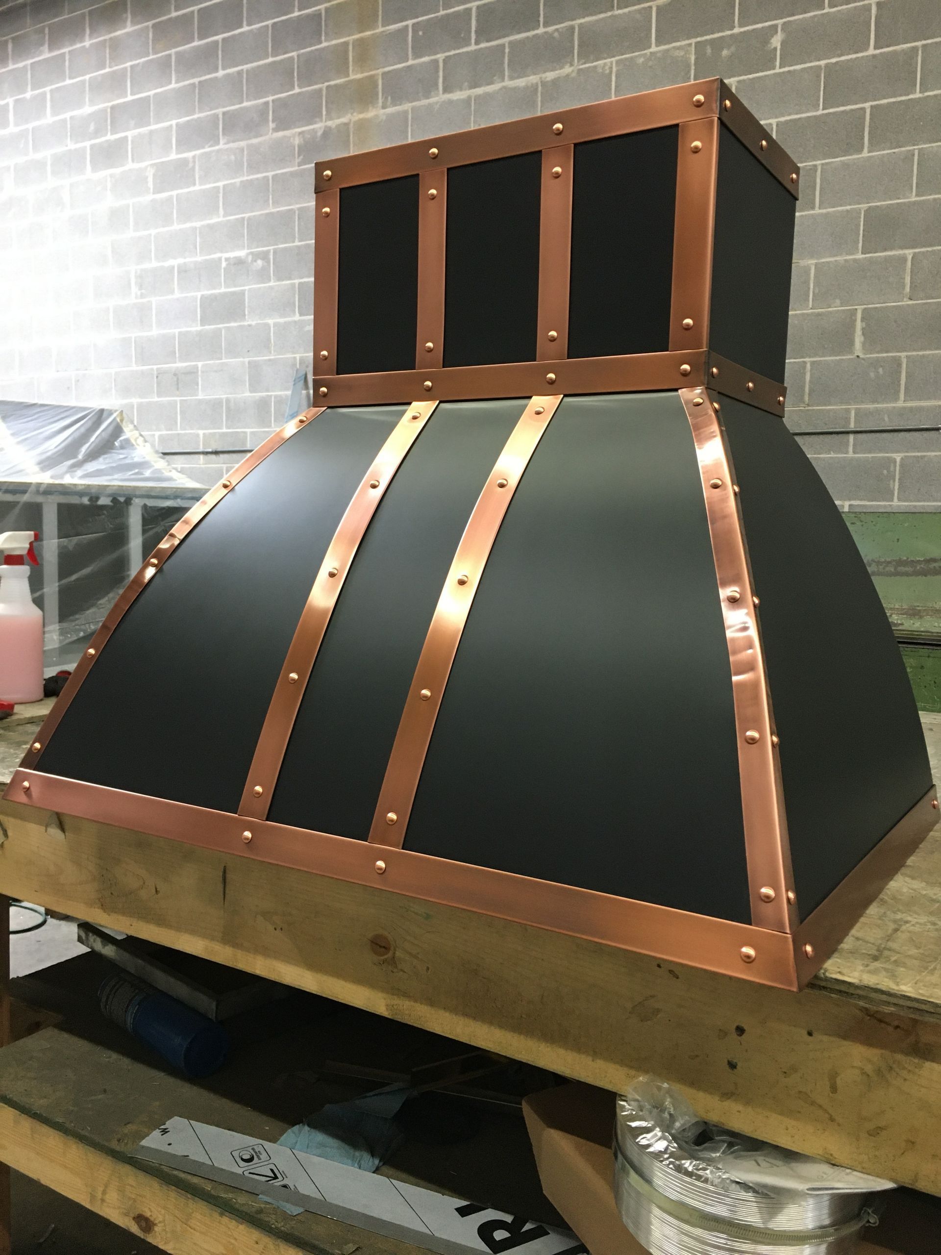 Large black metal hood with copper trim on a wooden workbench in a workshop