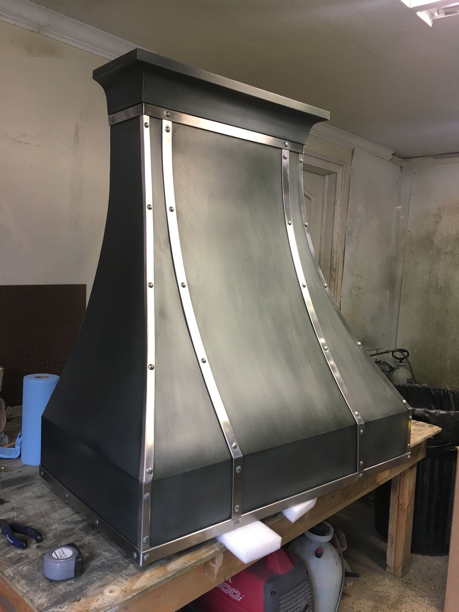 Large stainless steel and black metal hood or chimney unit in a workshop, mounted on a wooden bench.