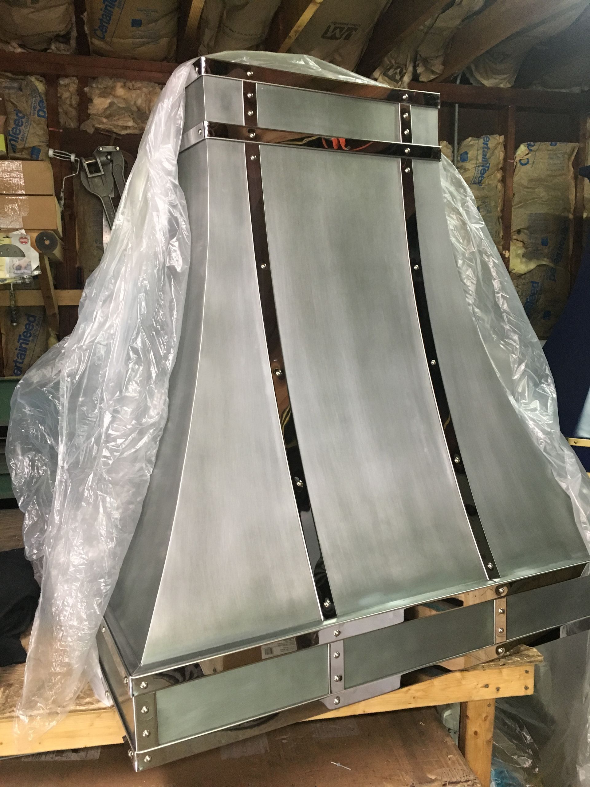 Large metal-framed object wrapped in clear plastic, leaning on a workbench in a workshop with insulation behind it