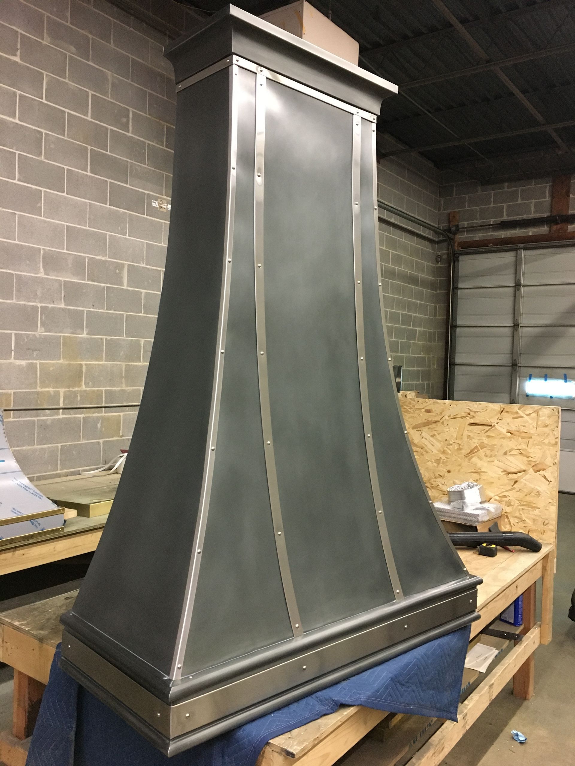Large metal ventilation hood in a workshop, with a gray brick wall and workbench in the background.