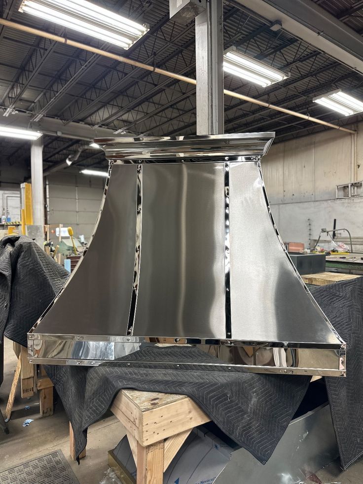 Shiny metal duct hood in a workshop, mounted on a worktable with tools and overhead lights.
