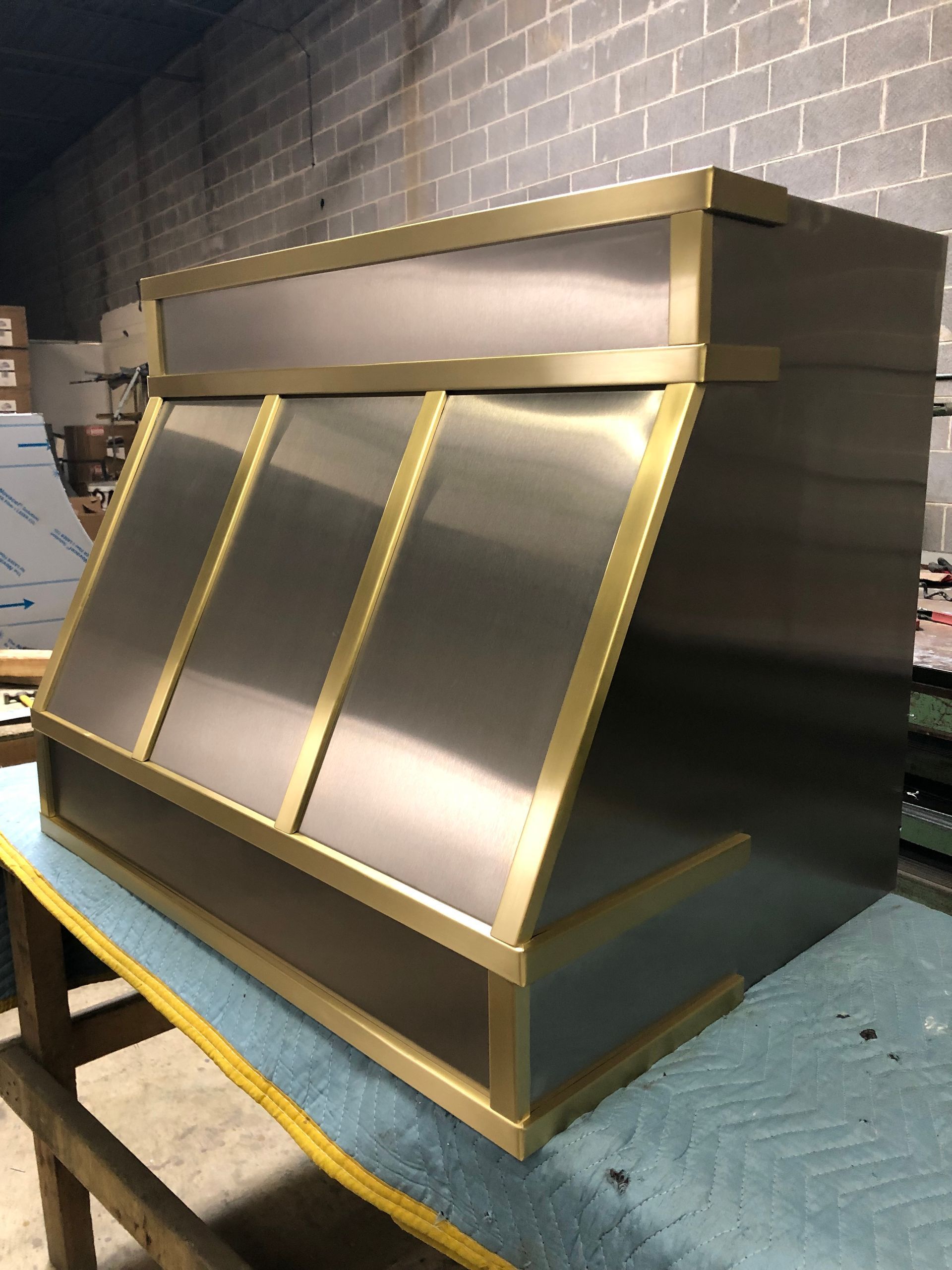 Metal industrial hopper or chute with gold trim sitting on a blue quilted pallet in a warehouse.